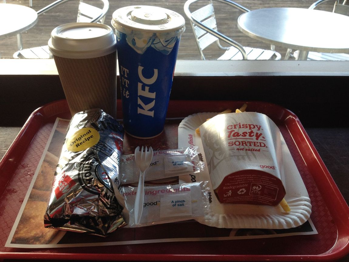 A fast-food meal on a tray: coffee, KFC drink, sandwich, wrap, cutlery, and sugar packets.