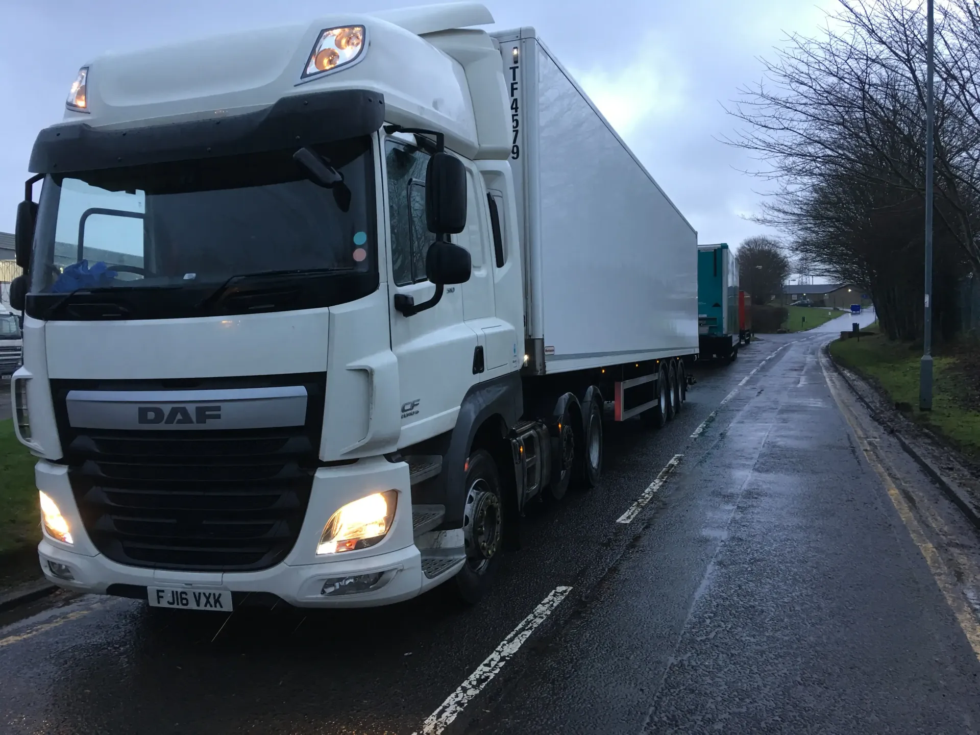 White DAF truck with a trailer on a wet road, parked with other vehicles in the background. Overcast sky.