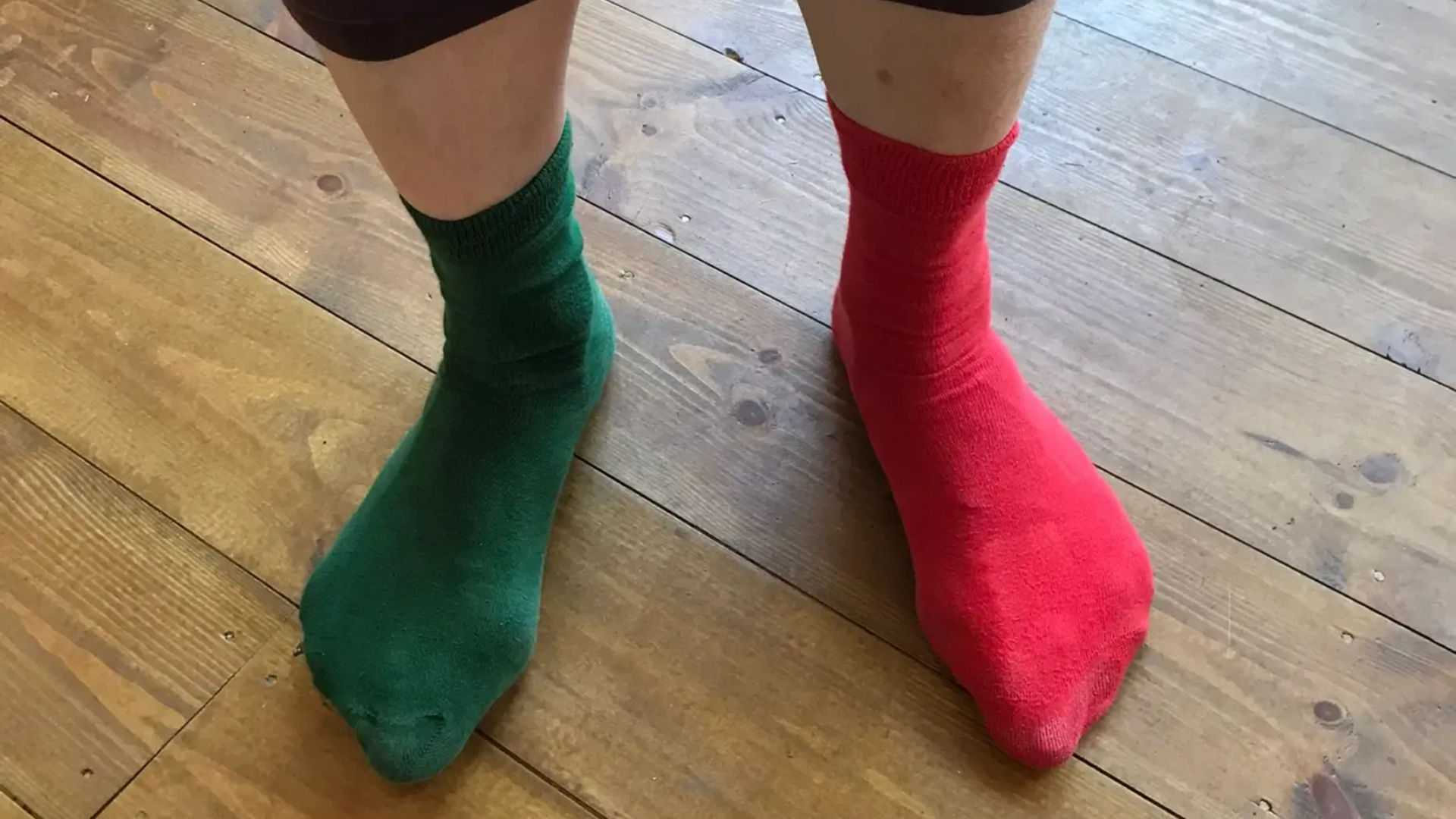 Pair of feet wearing mismatched green and red socks on a wooden floor.