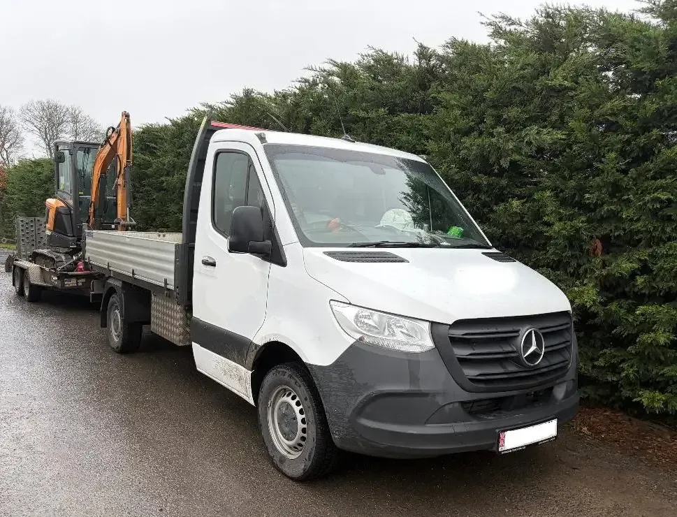 White Mercedes truck with a flatbed pulling a trailer carrying a small excavator on a road.