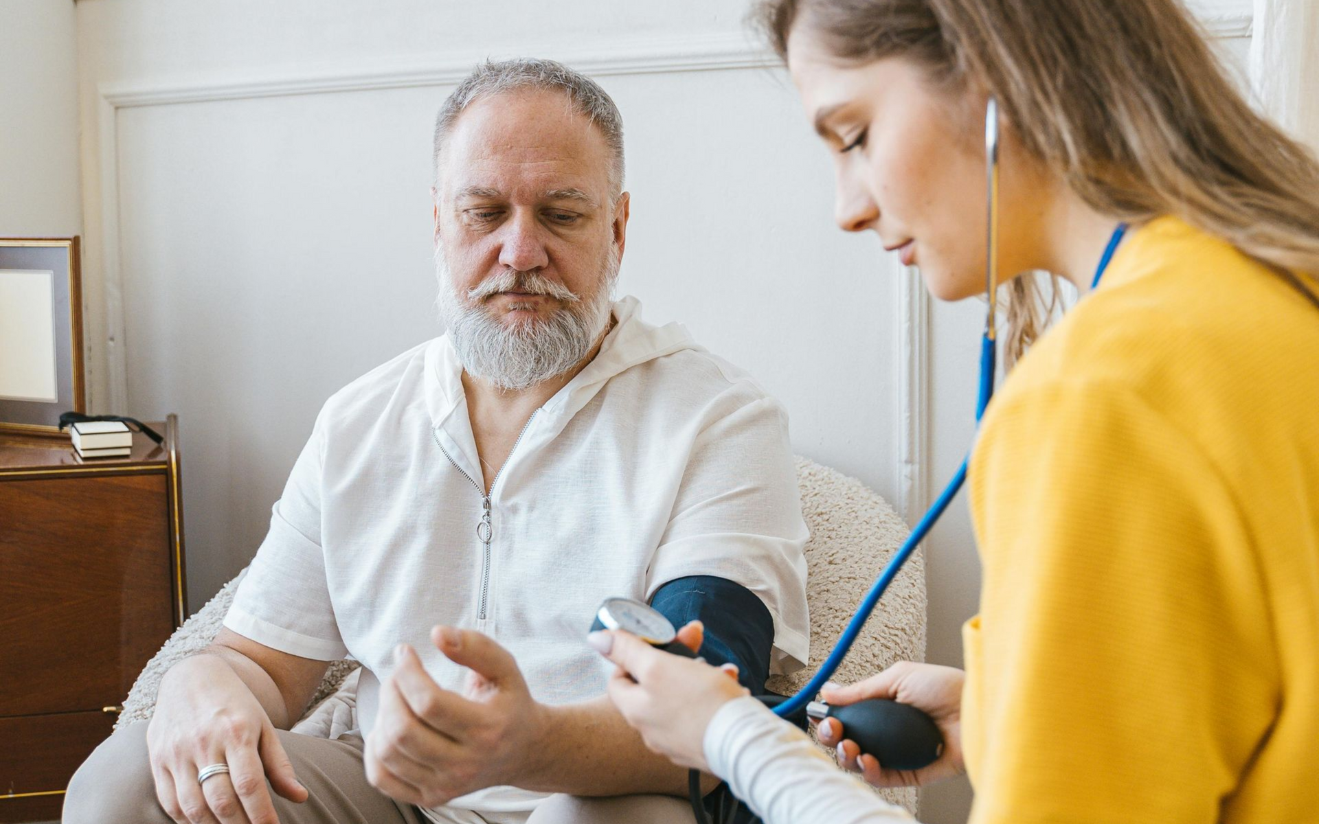 A healthcare worker taking a patient's blood pressure with a stethoscope in a home setting.