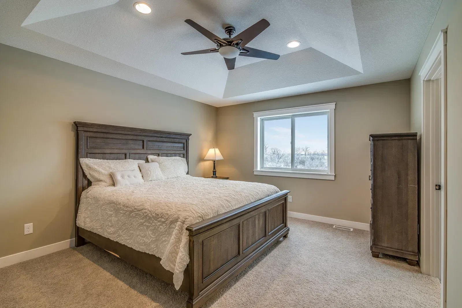 Bedroom with a large bed, nightstand, dresser, and a window. Beige walls and carpet. Ceiling fan.