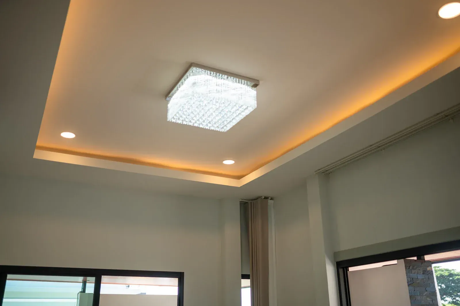 Ceiling with recessed lighting and a crystal chandelier in a room with windows and curtains.