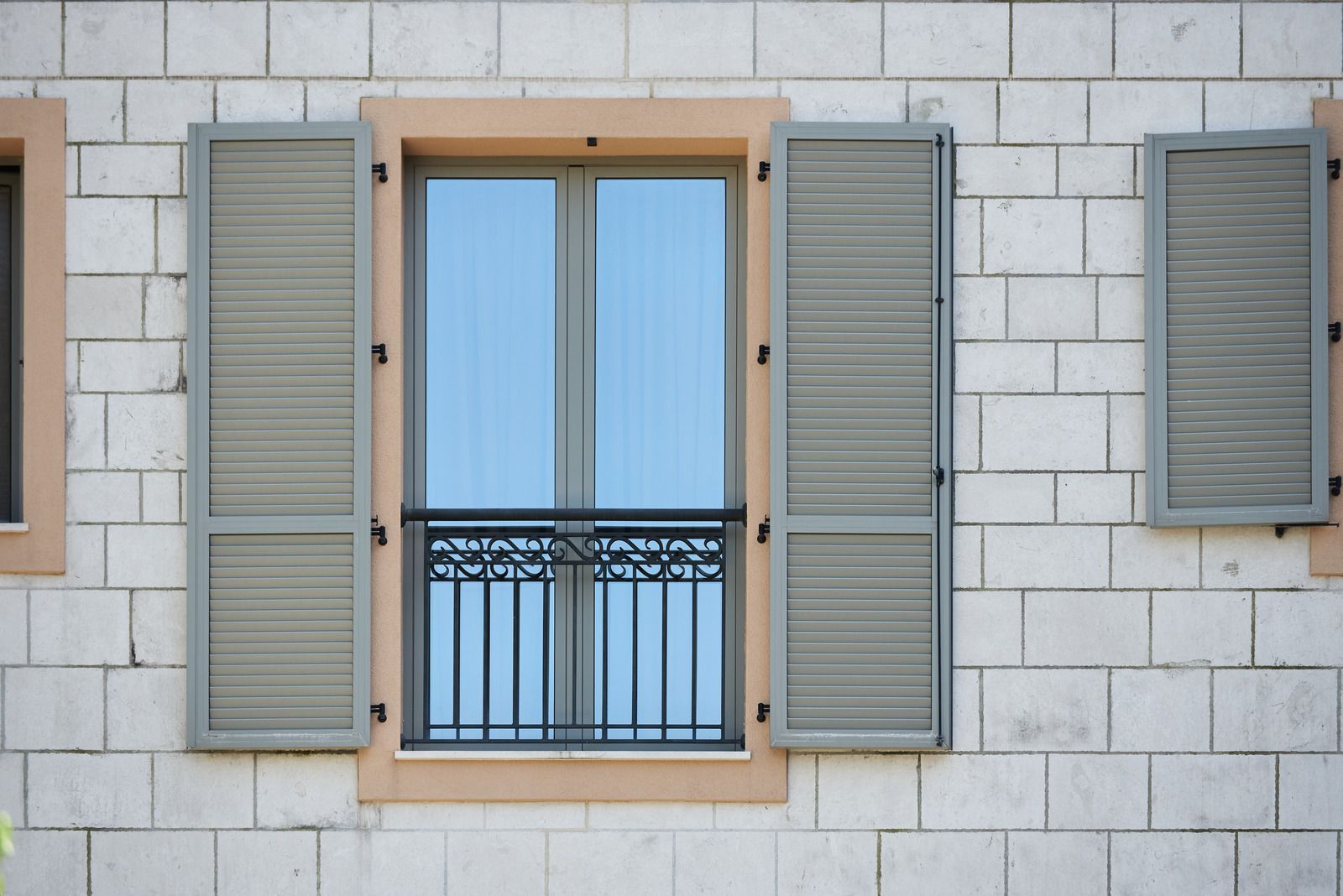Window with open gray shutters, black wrought-iron railing, and light blue sky reflection on a white brick wall.