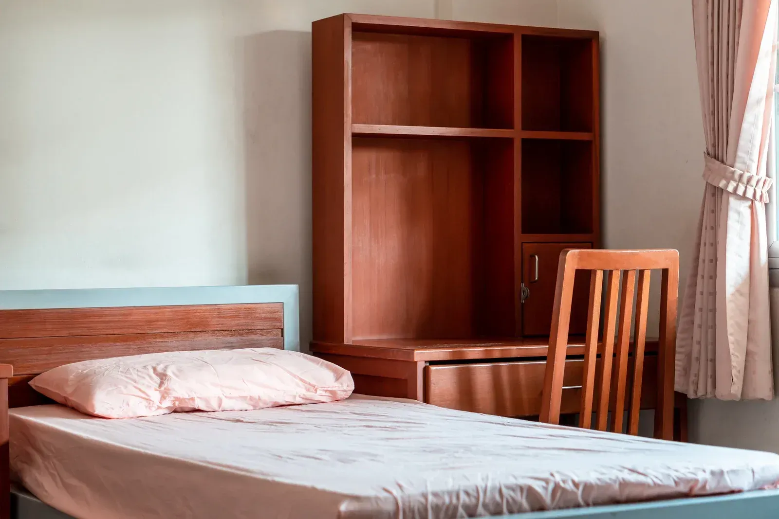 A simple bedroom with a single bed, wooden bookcase, chair, and curtained window.