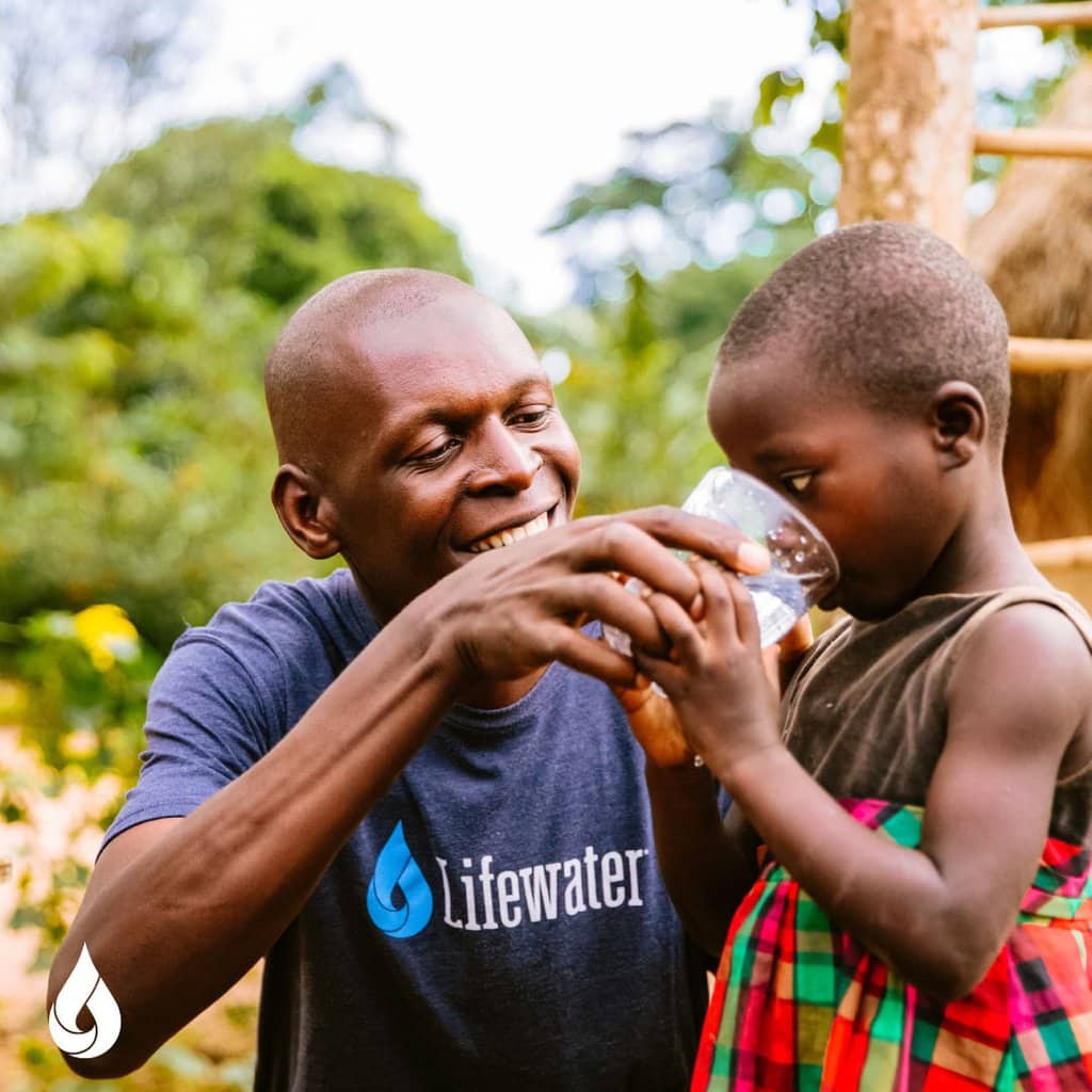 A man wearing a lifewater shirt is giving a little girl a glass of water