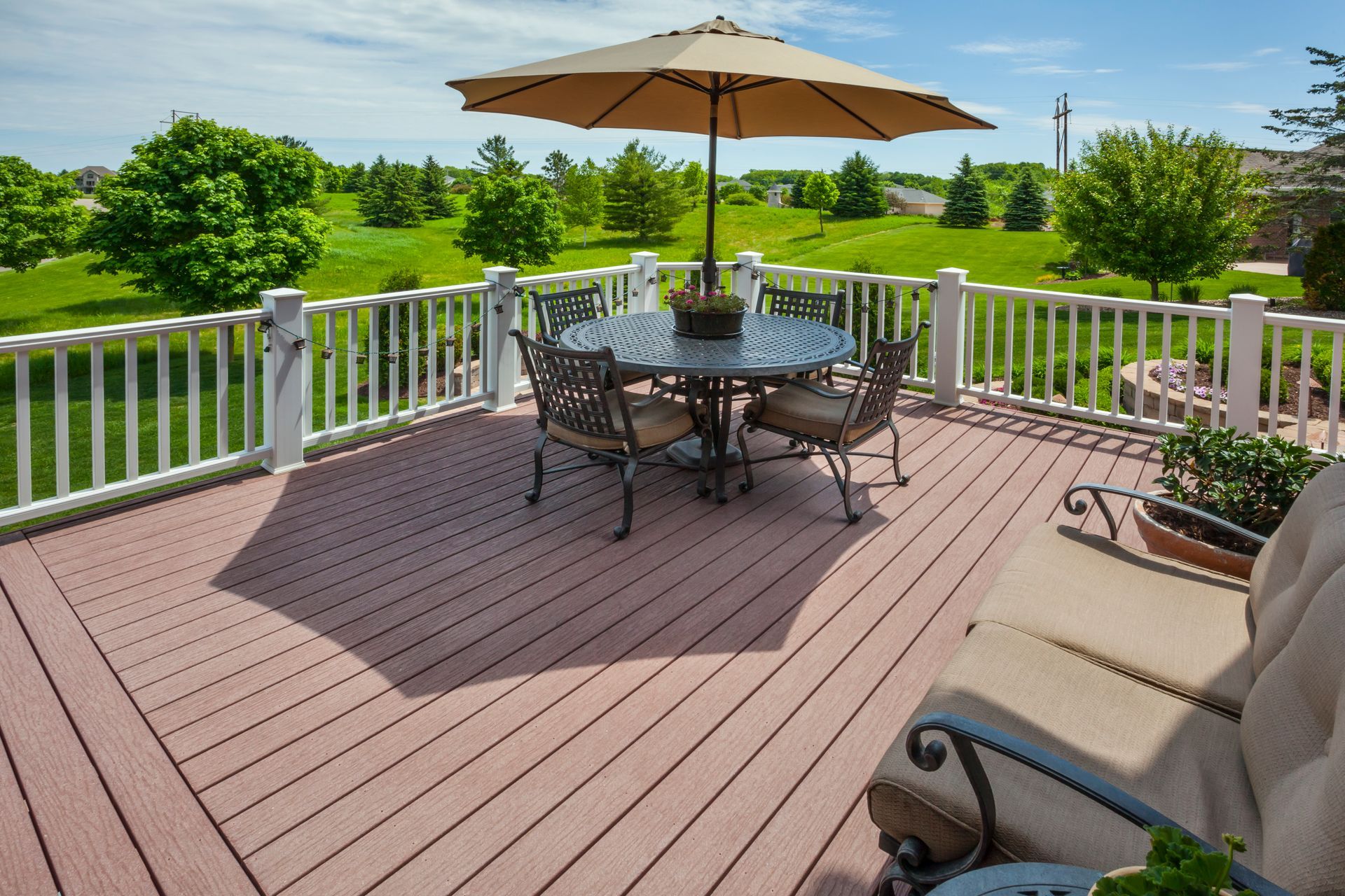 Wooden deck with round table, chairs, and umbrella overlooking green landscape on a sunny day.