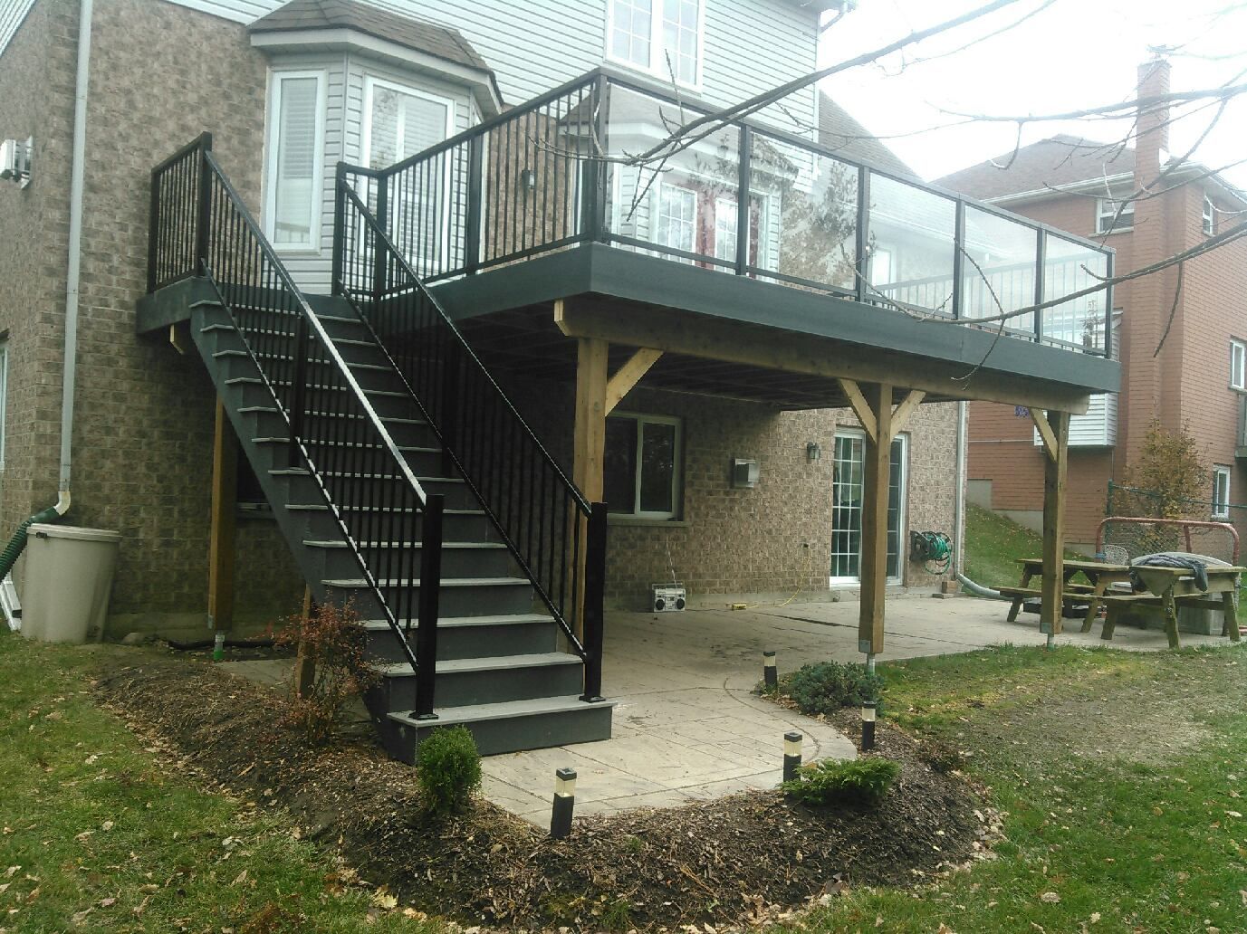 Backyard deck with stairs, glass railing, black railing, and ground-level patio.