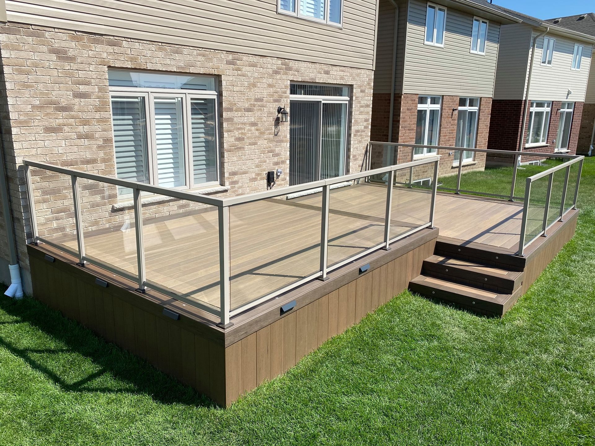 Composite deck with glass railing, steps, and house backdrop on a grassy lawn.
