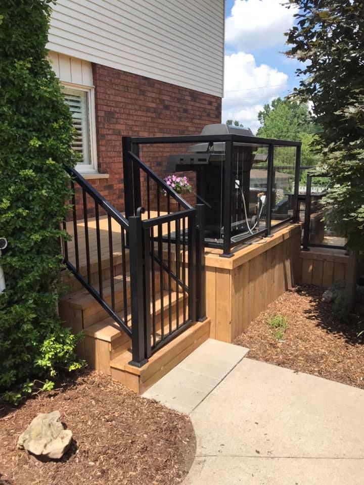 Wooden deck with black railing and glass panels, a grill, and a brick house. Sunny day.