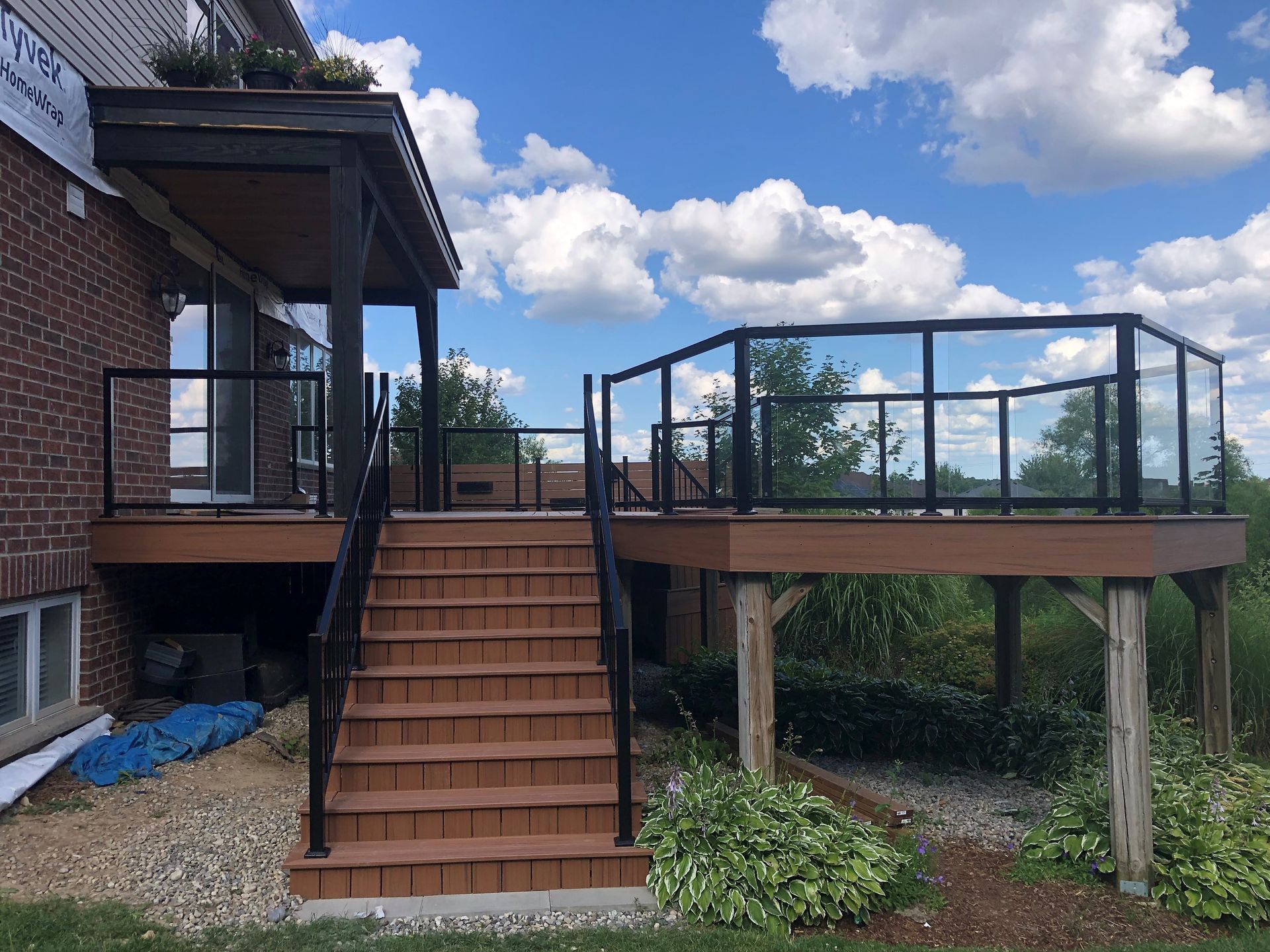 Wooden deck with glass railing and stairs attached to a brick house on a sunny day.