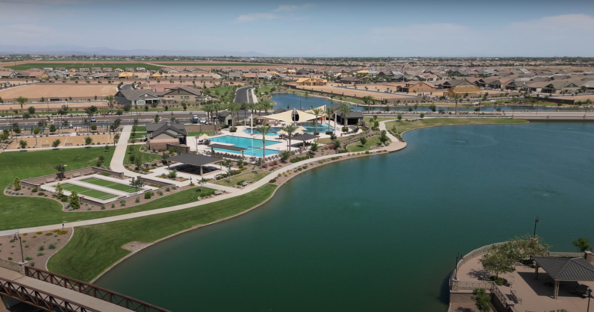 Aerial view of a lakefront community with swimming pools, houses, and a bridge, under a clear blue sky.
