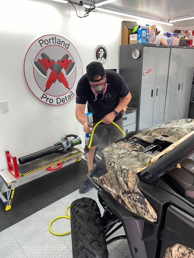 Man cleaning a camouflage car hood with a hose in a garage, with lockers and tools nearby.