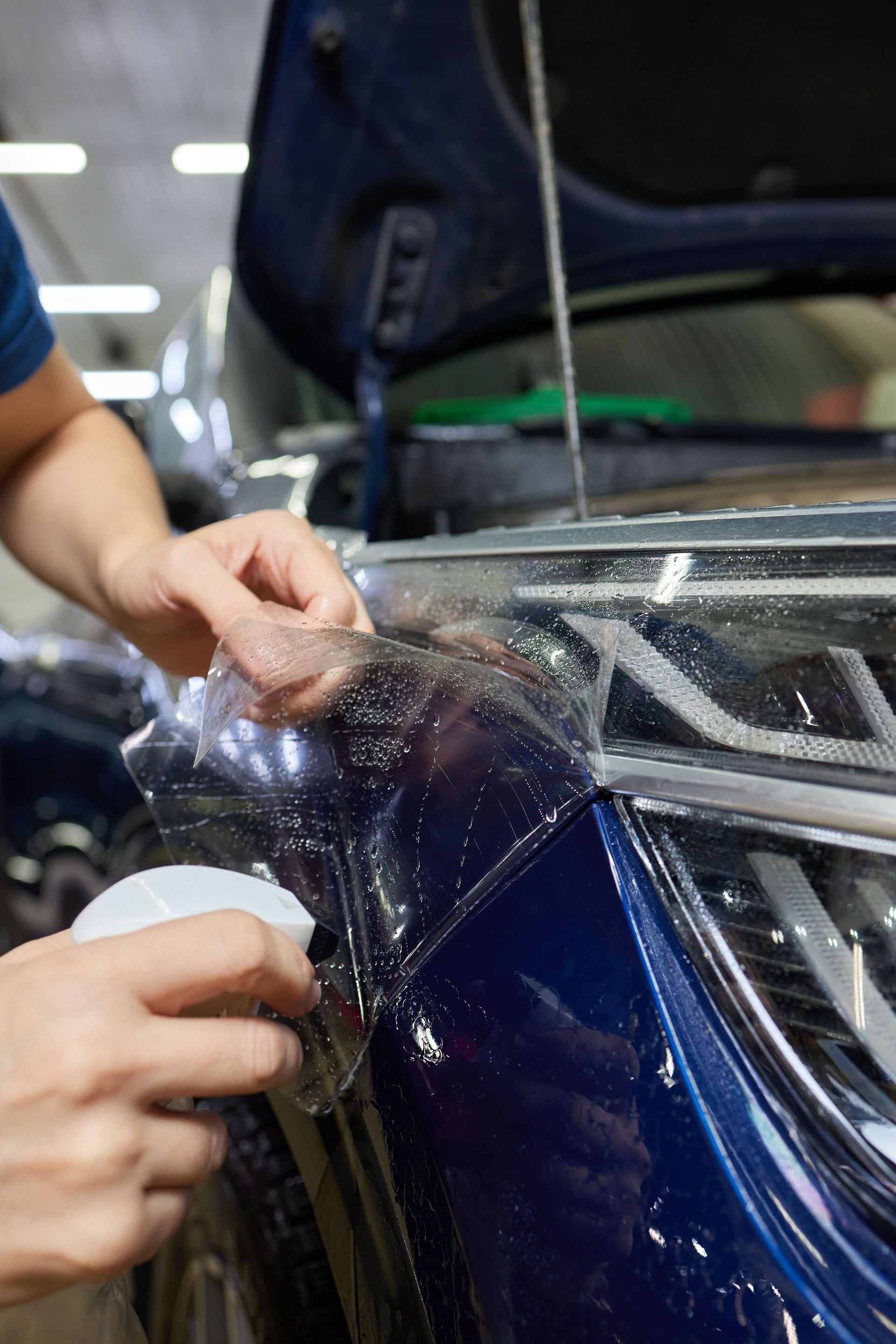 Hands applying clear protective film to a dark blue car headlight.