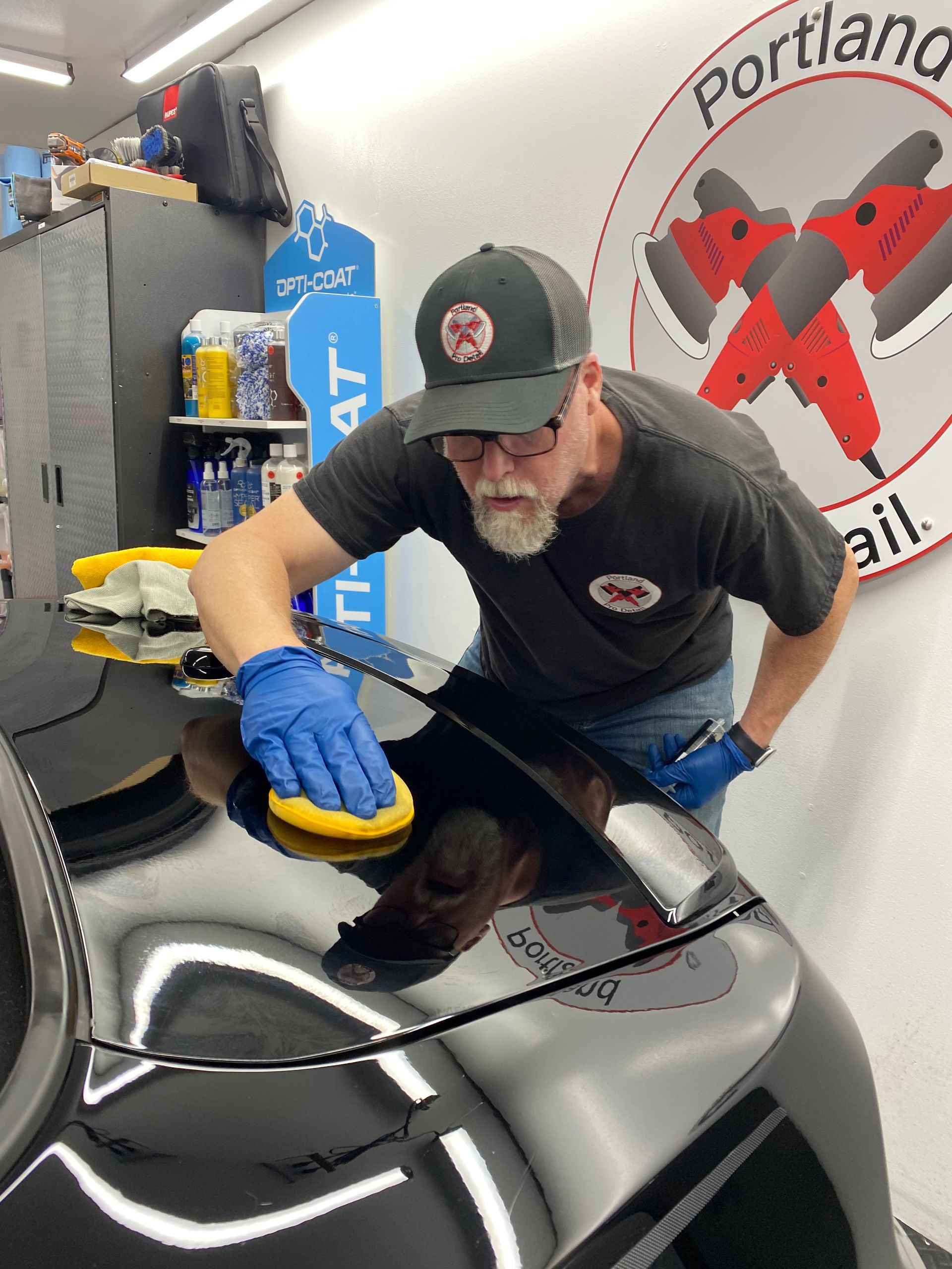 Man polishing a black car hood with a blue sponge in an auto detailing shop