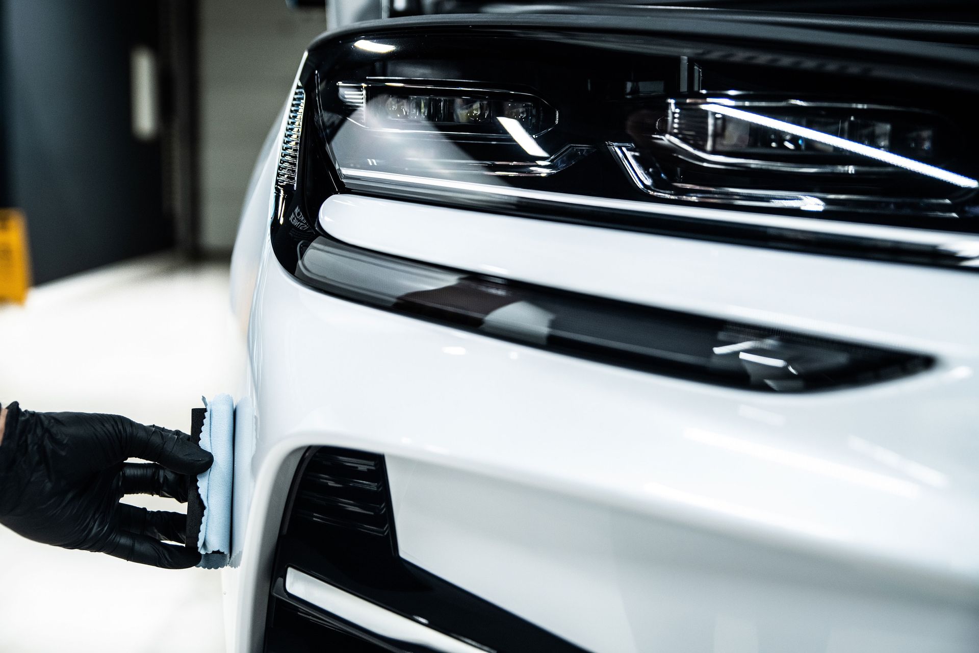 Gloved hand wiping the front bumper of a white car in a garage
