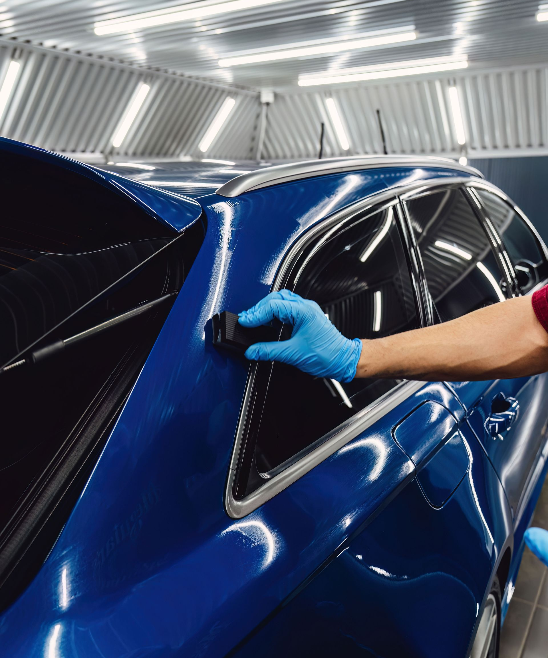 Blue car body being polished by a gloved hand in a bright workshop with overhead lights