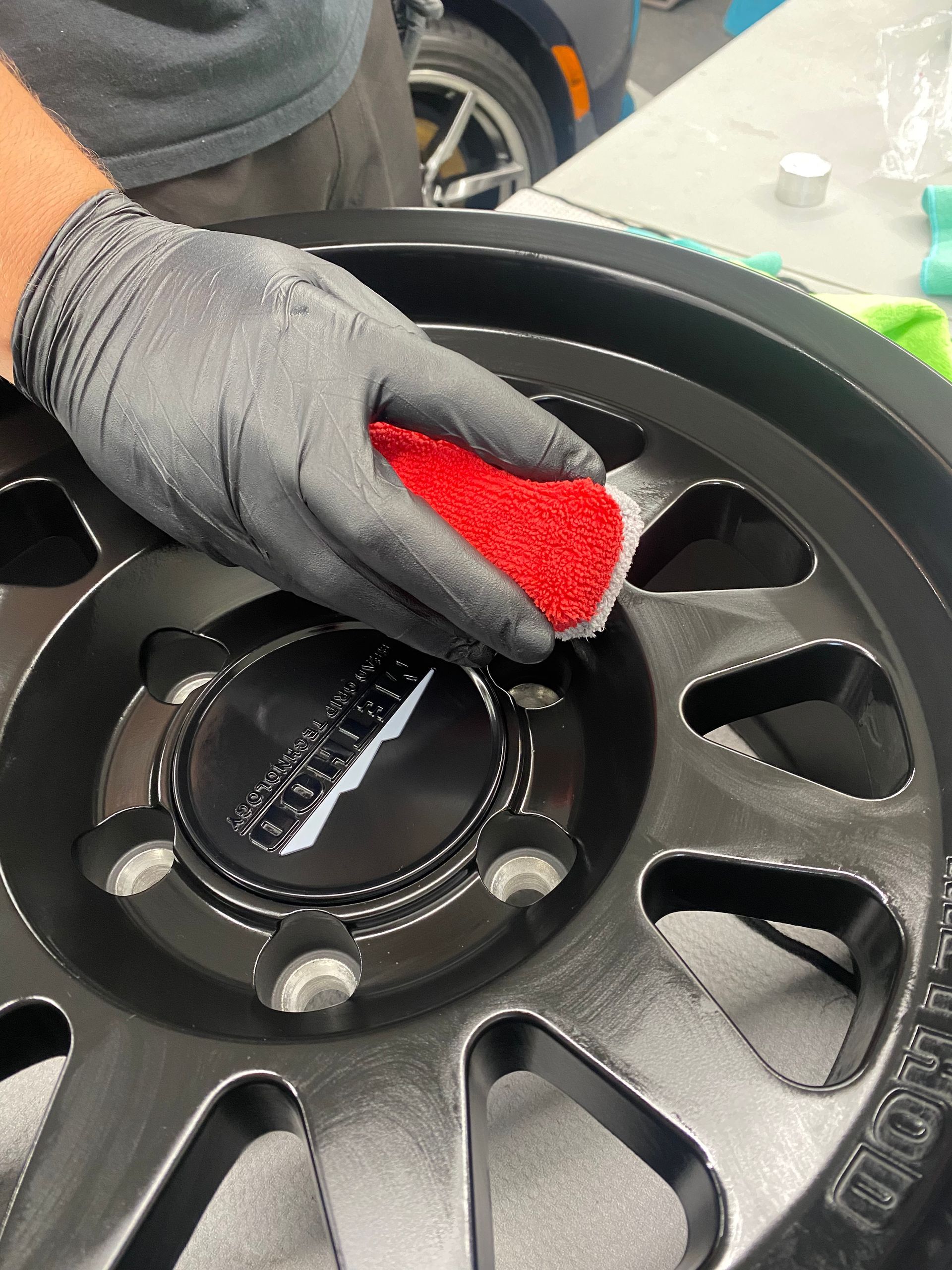 Gloved hand wiping a black car wheel rim with a red applicator pad