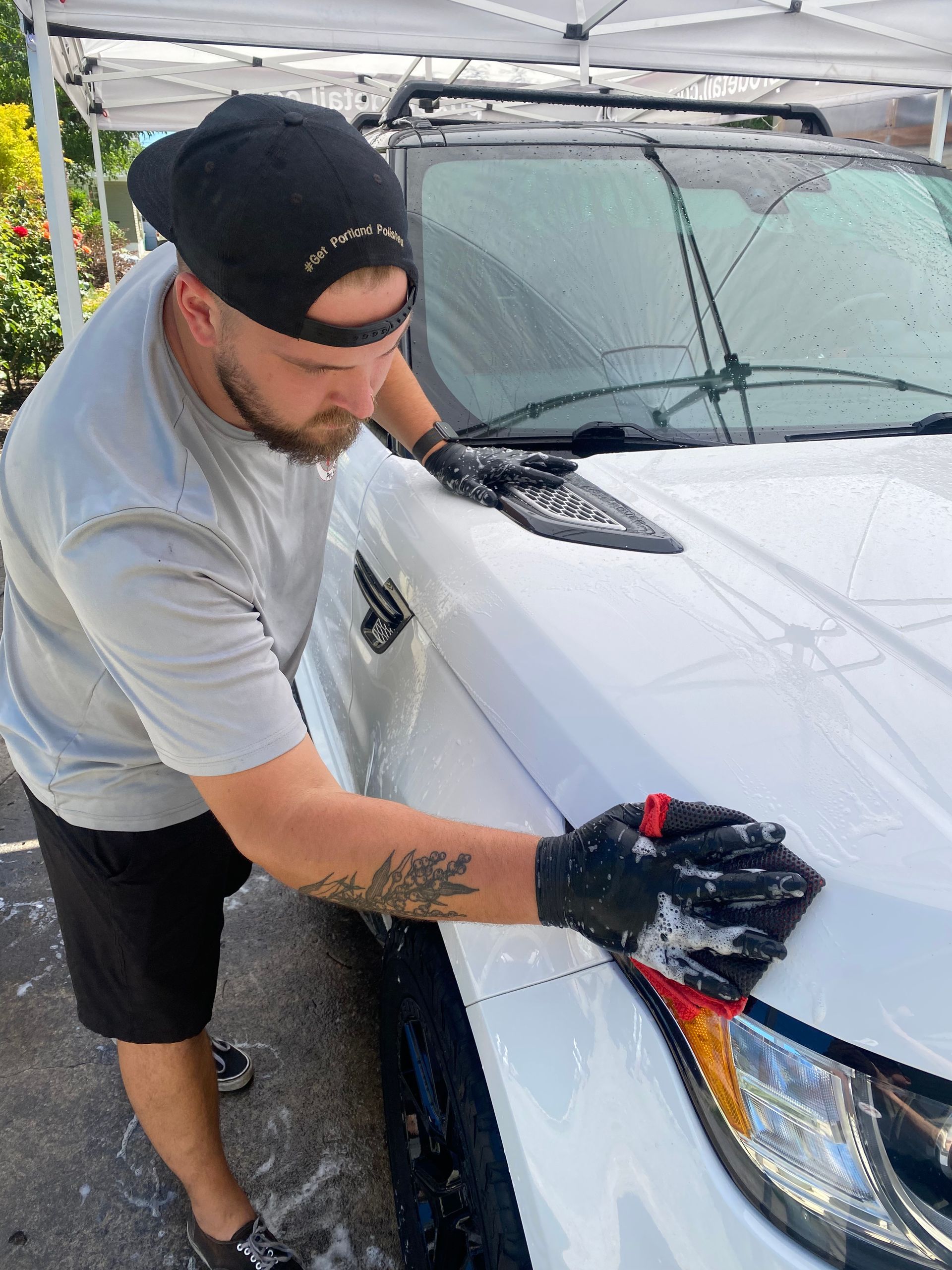 Person washing a white SUV with a sponge in a driveway under a canopy