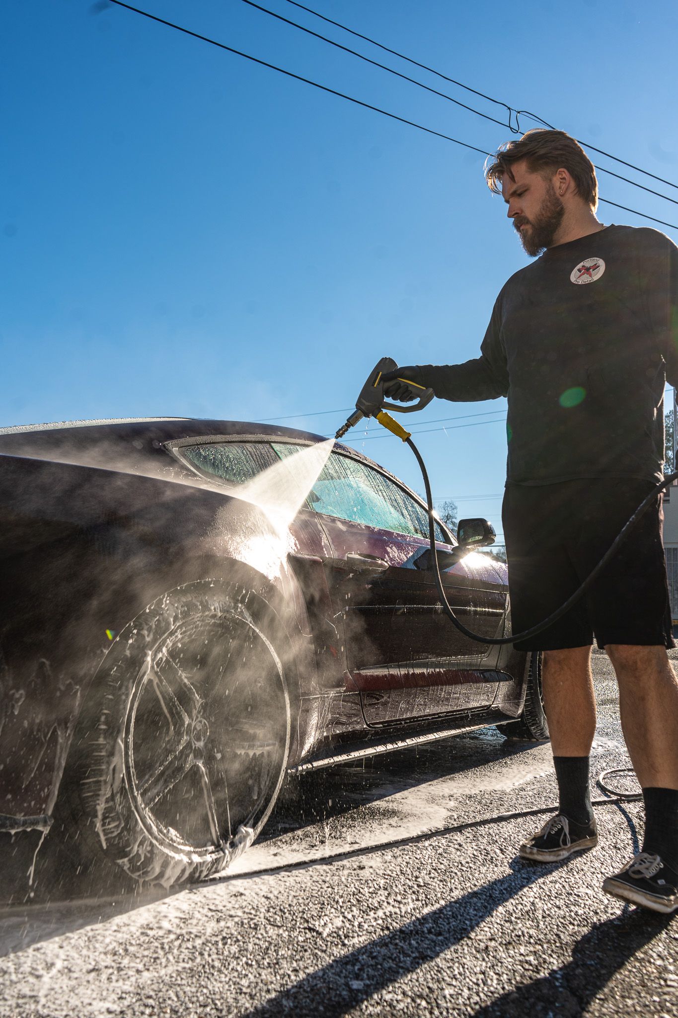 Person washing a black car outdoors with a pressure washer on a sunny day