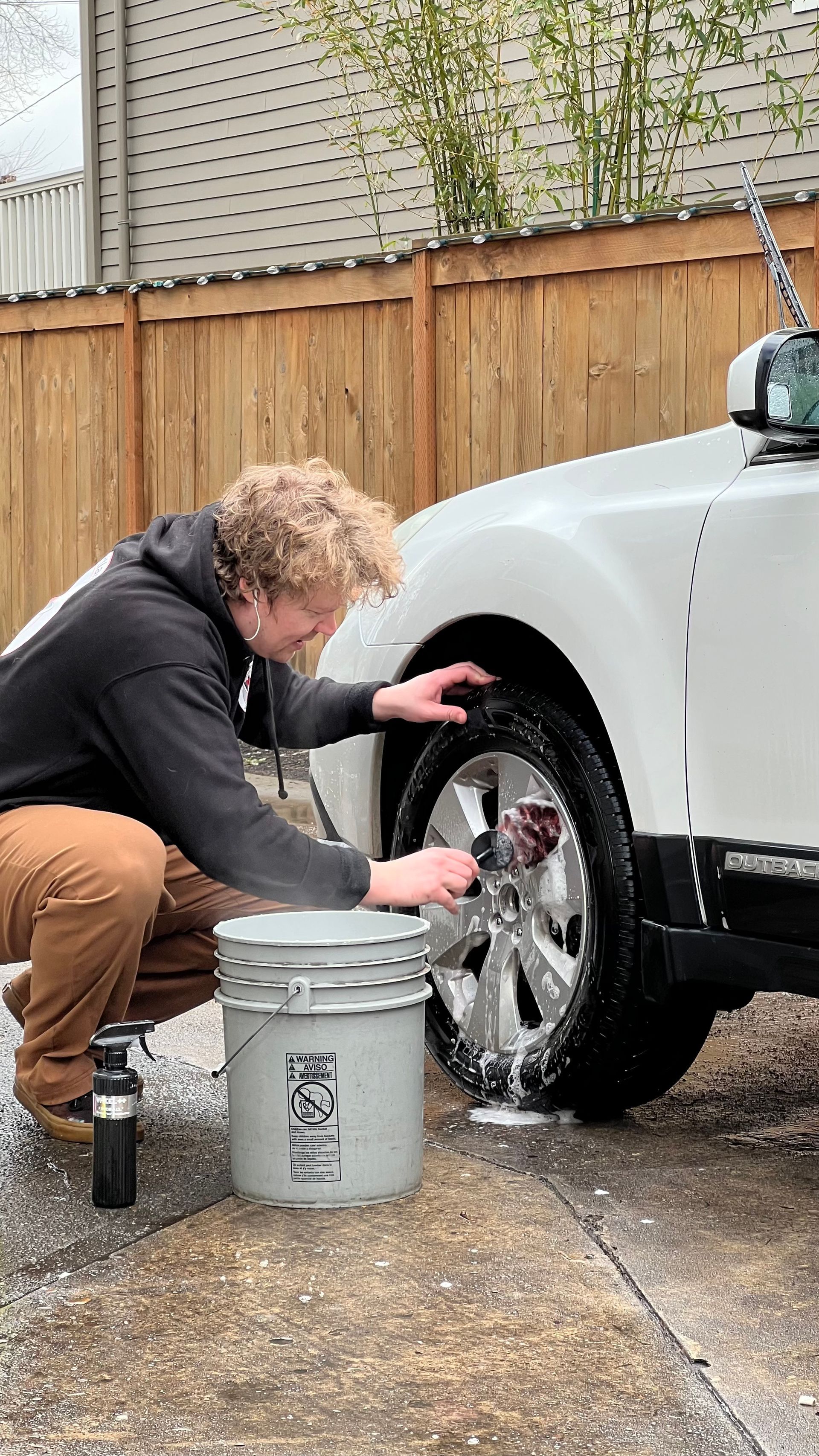 Person washing a white car wheel with a brush and bucket in a driveway