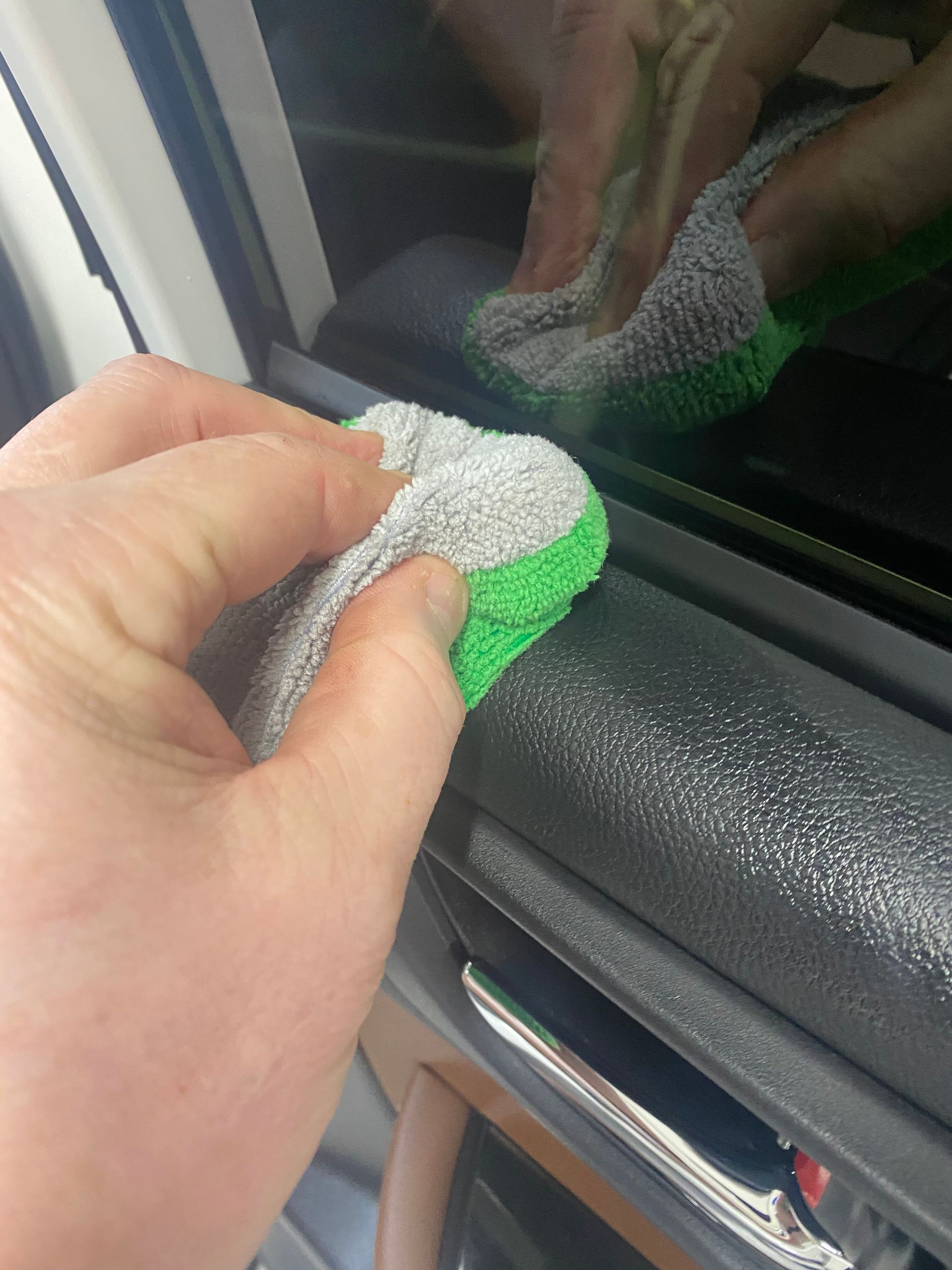 Hand wiping a car window with a gray and green sponge, reflected in the glass