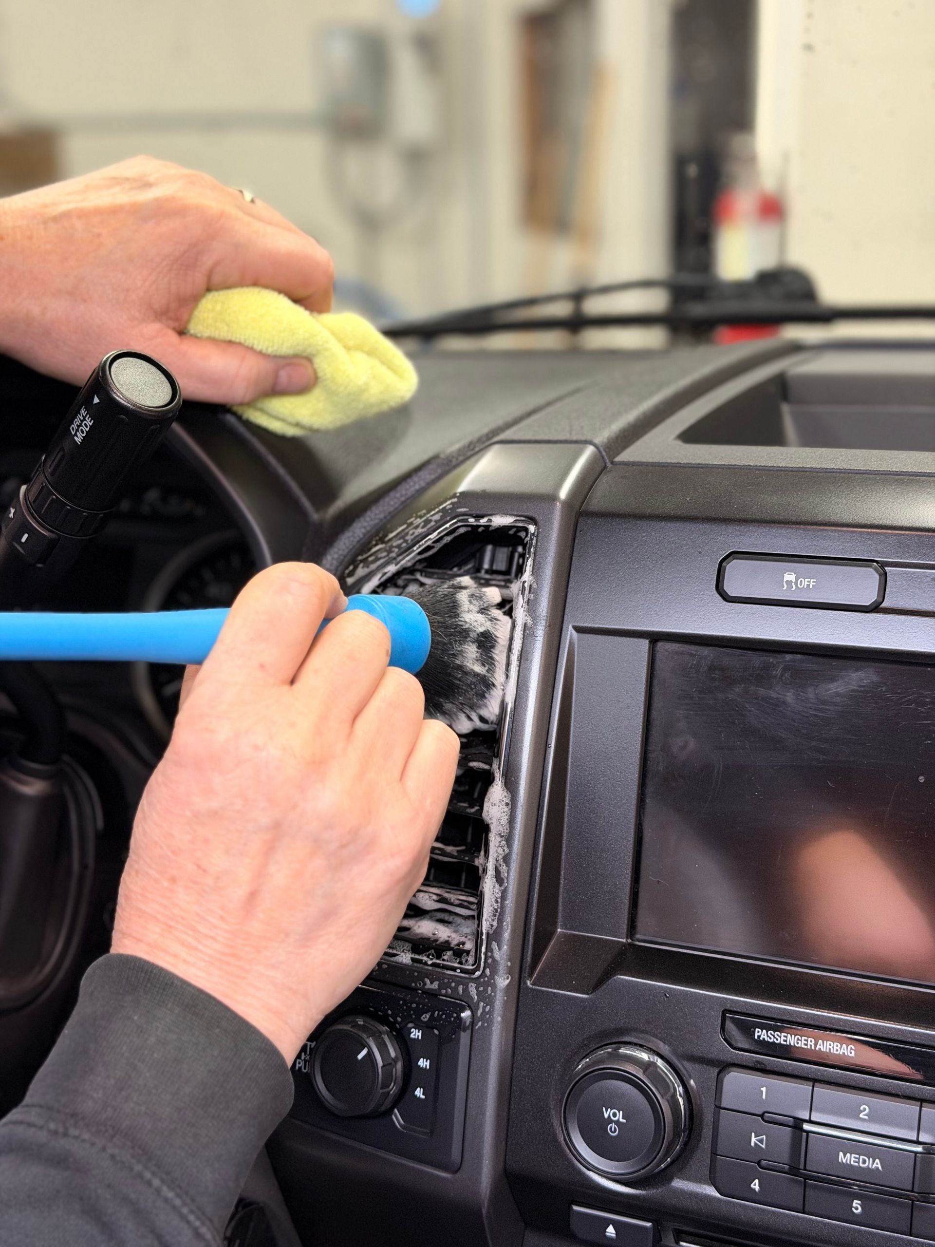 Hands cleaning dust from a car dashboard with a blue brush and cloth