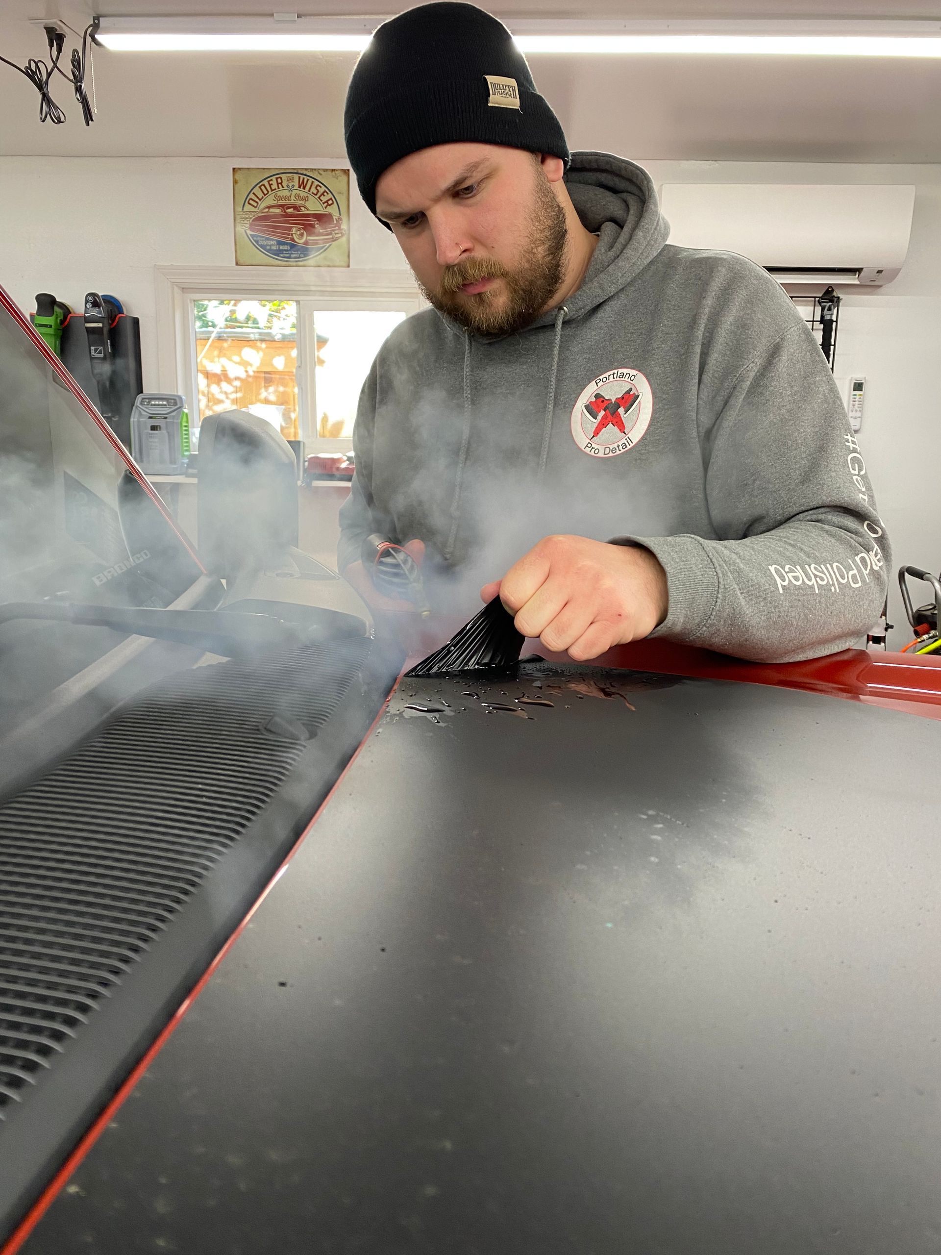 Man in gray hoodie and black beanie cleaning a black tabletop with a cloth in a garage or workshop.