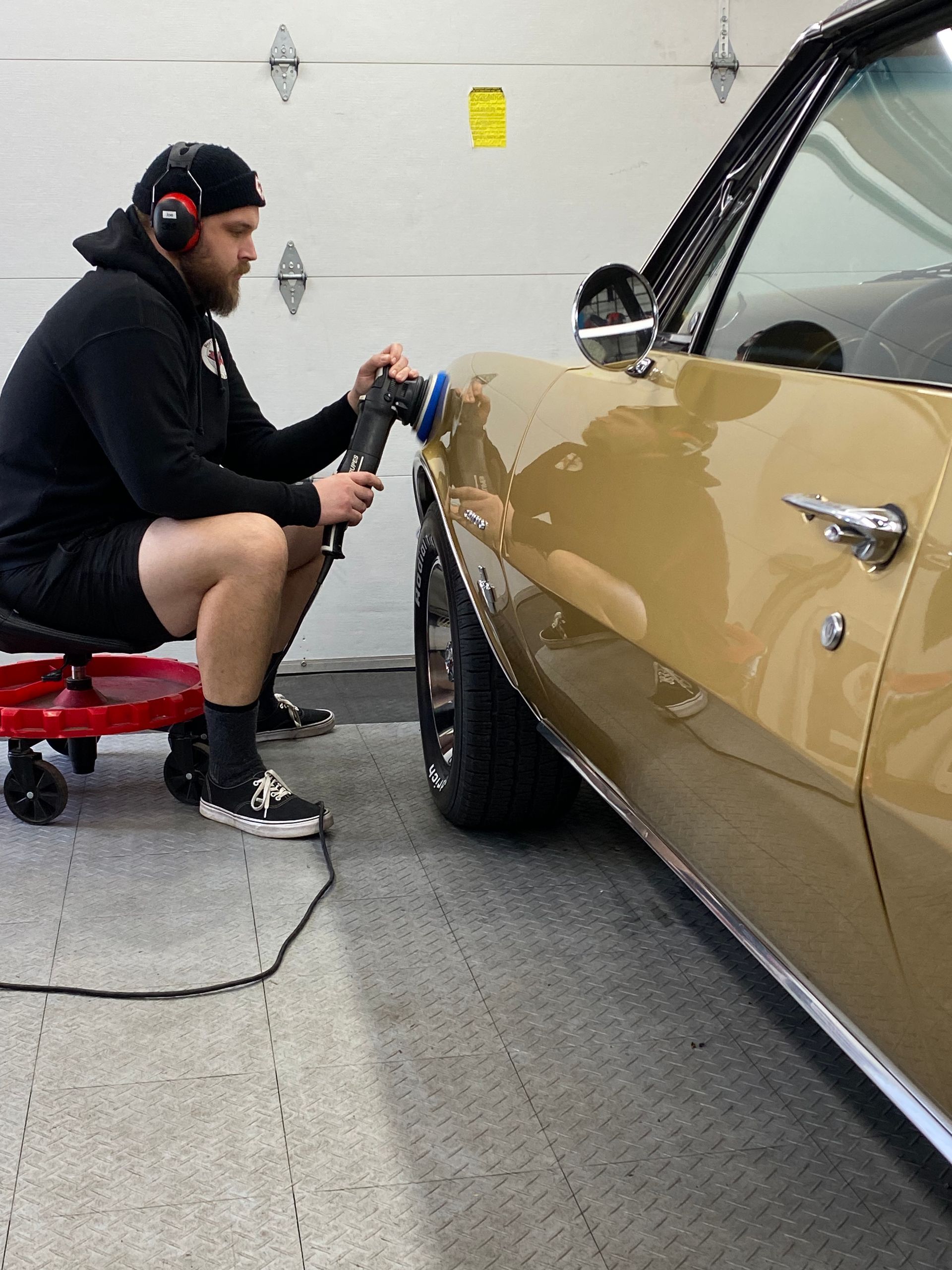 A worker polishing a tan car in a garage with a handheld buffer.