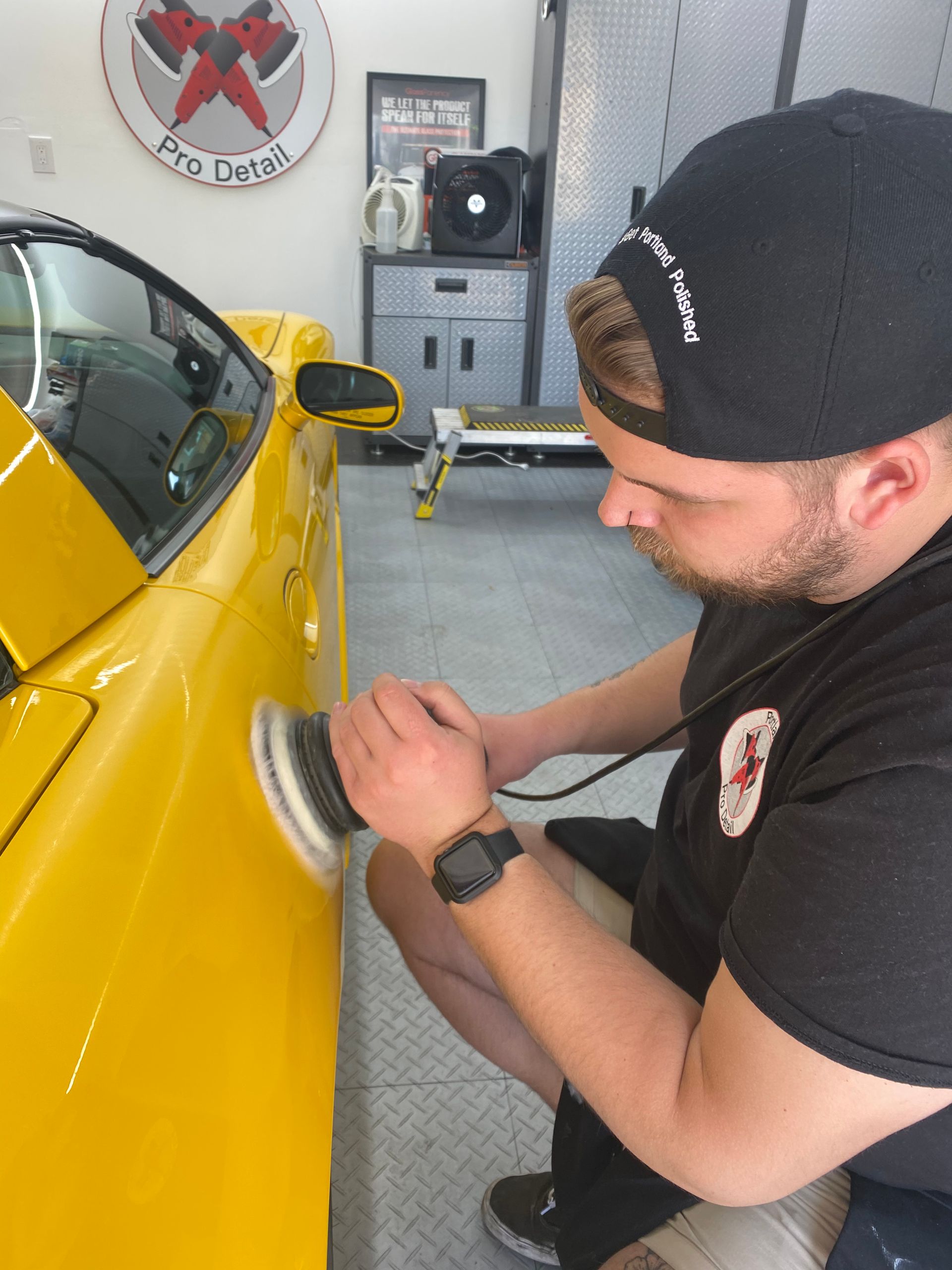 Man polishing a yellow car door in a garage with a buffer tool