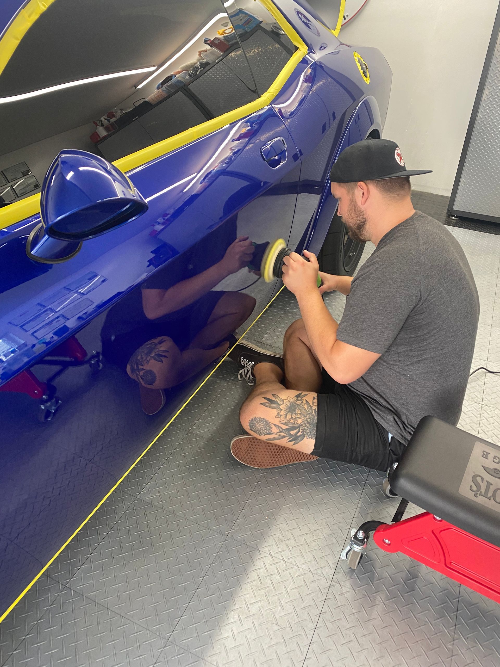 Man polishing a blue car door in a garage, with yellow tape outlining the vehicle.