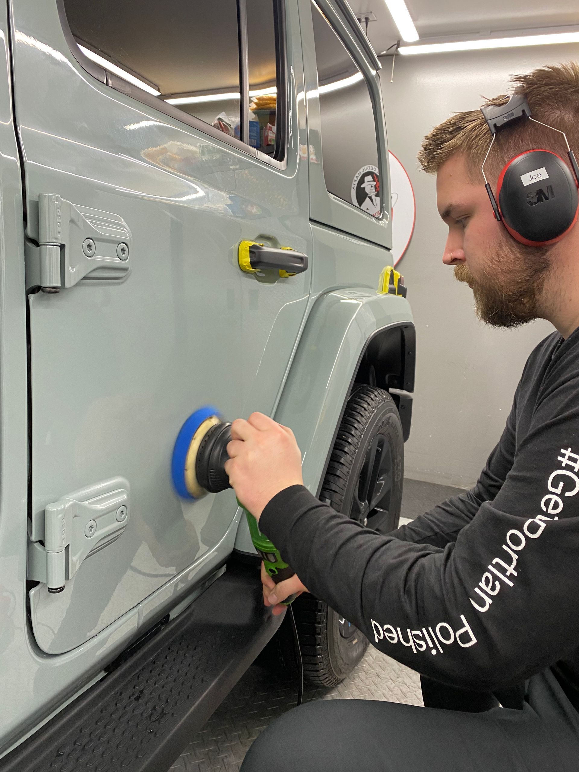Man in headphones polishing a gray vehicle with a handheld buffer in a workshop