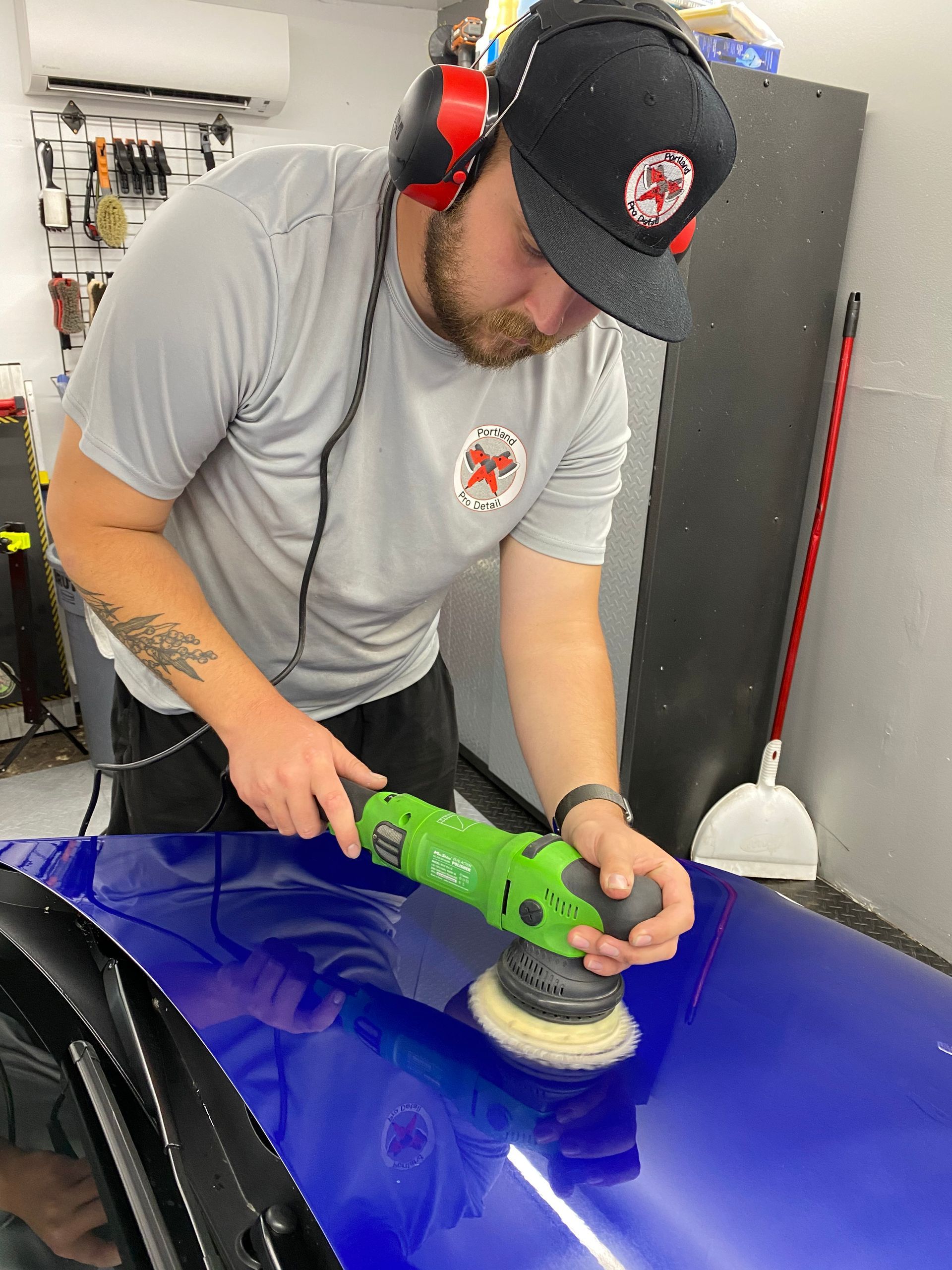 Man polishing a blue car panel with a green buffer in a garage, wearing headphones and a cap