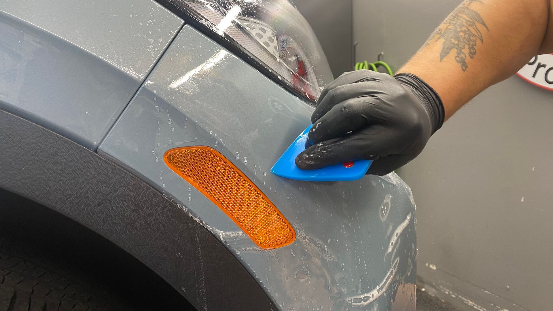 Gloved hand polishing a gray car fender with a blue applicator pad near an orange reflector.