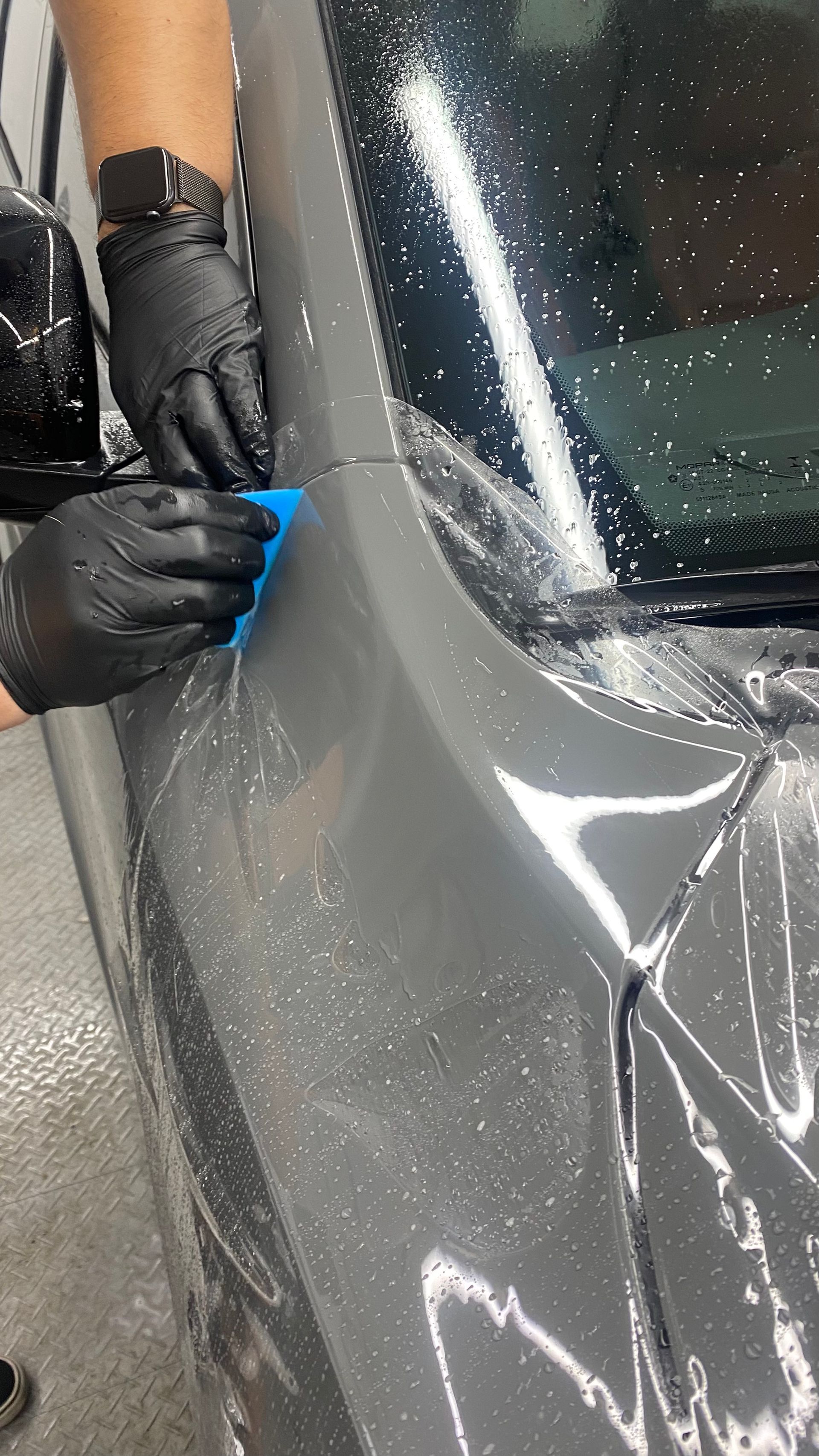 Gloved hands applying blue tape to a car’s windshield edge beside a clear protective film