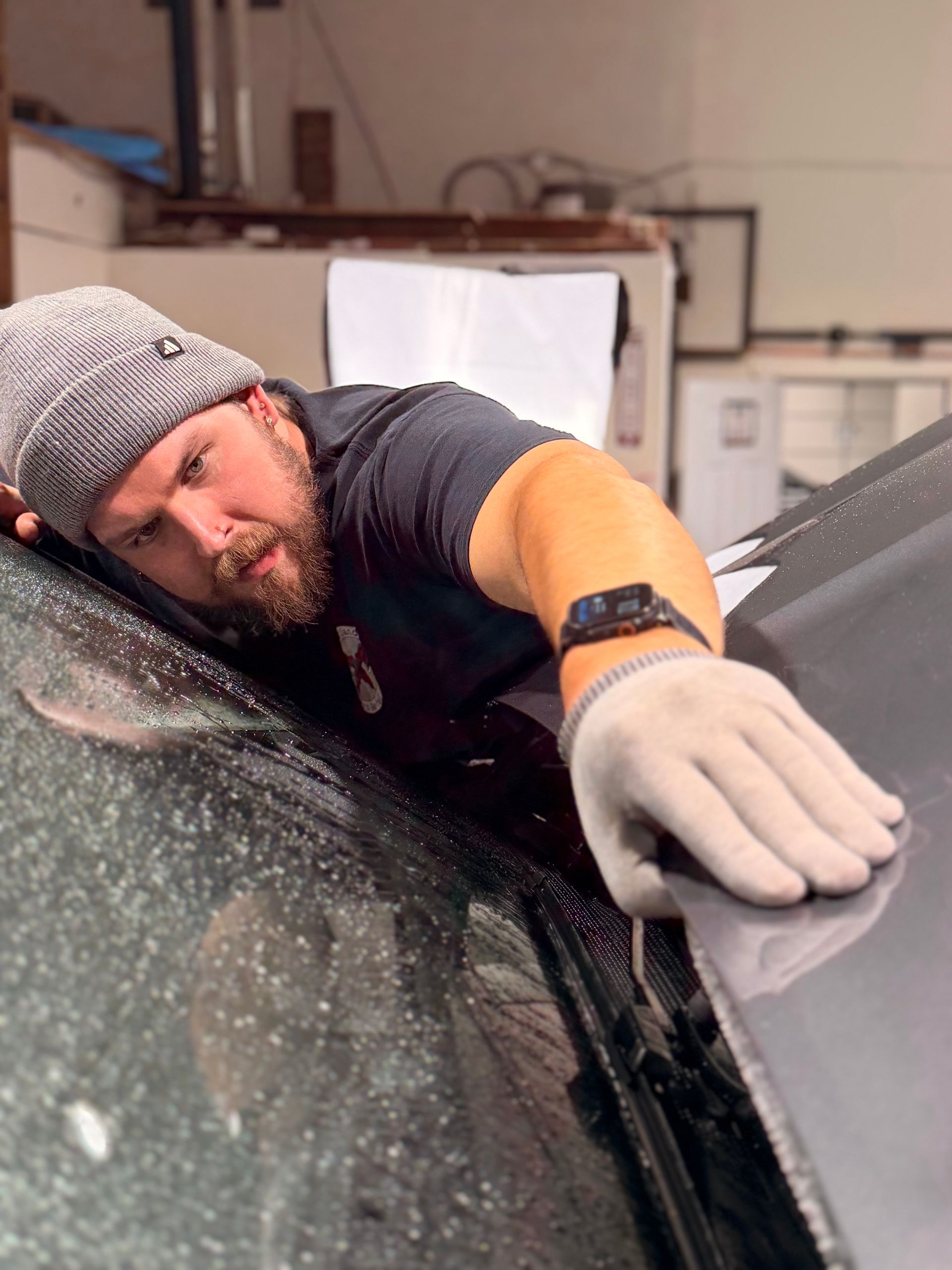 Worker wiping a car windshield in a garage, leaning over the hood with a gloved hand.