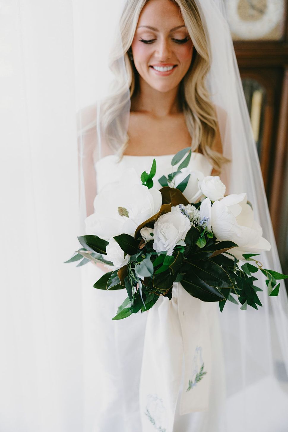 Bride in a white veil holding a bouquet of white flowers and greenery, smiling indoors
