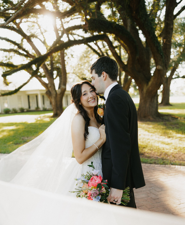 Bride and groom smiling outdoors beneath a large tree, holding a bouquet and wearing wedding attire
