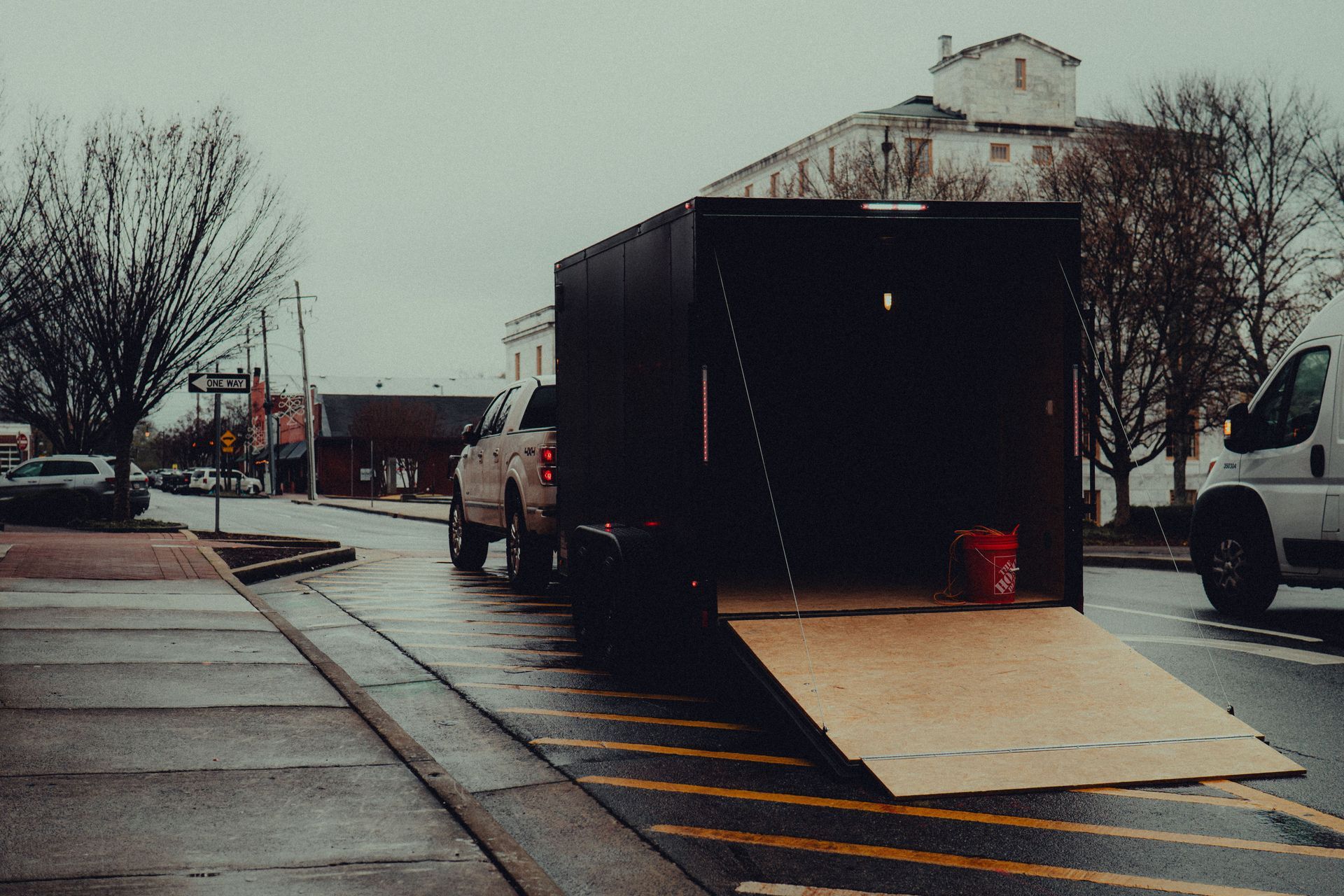 Person unloading a black trailer on a wet street, with a ramp down and a white truck nearby.