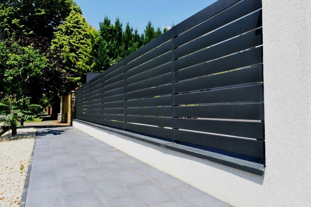 Modern black slatted fence along a paved walkway beside a white wall and green trees