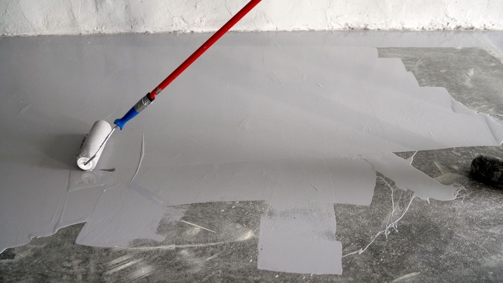 Person rolling gray paint on a floor with a white paint roller attached to a red handle