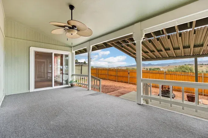 Covered patio with carpet, ceiling fan, sliding door, and views of fenced backyard and open fields.