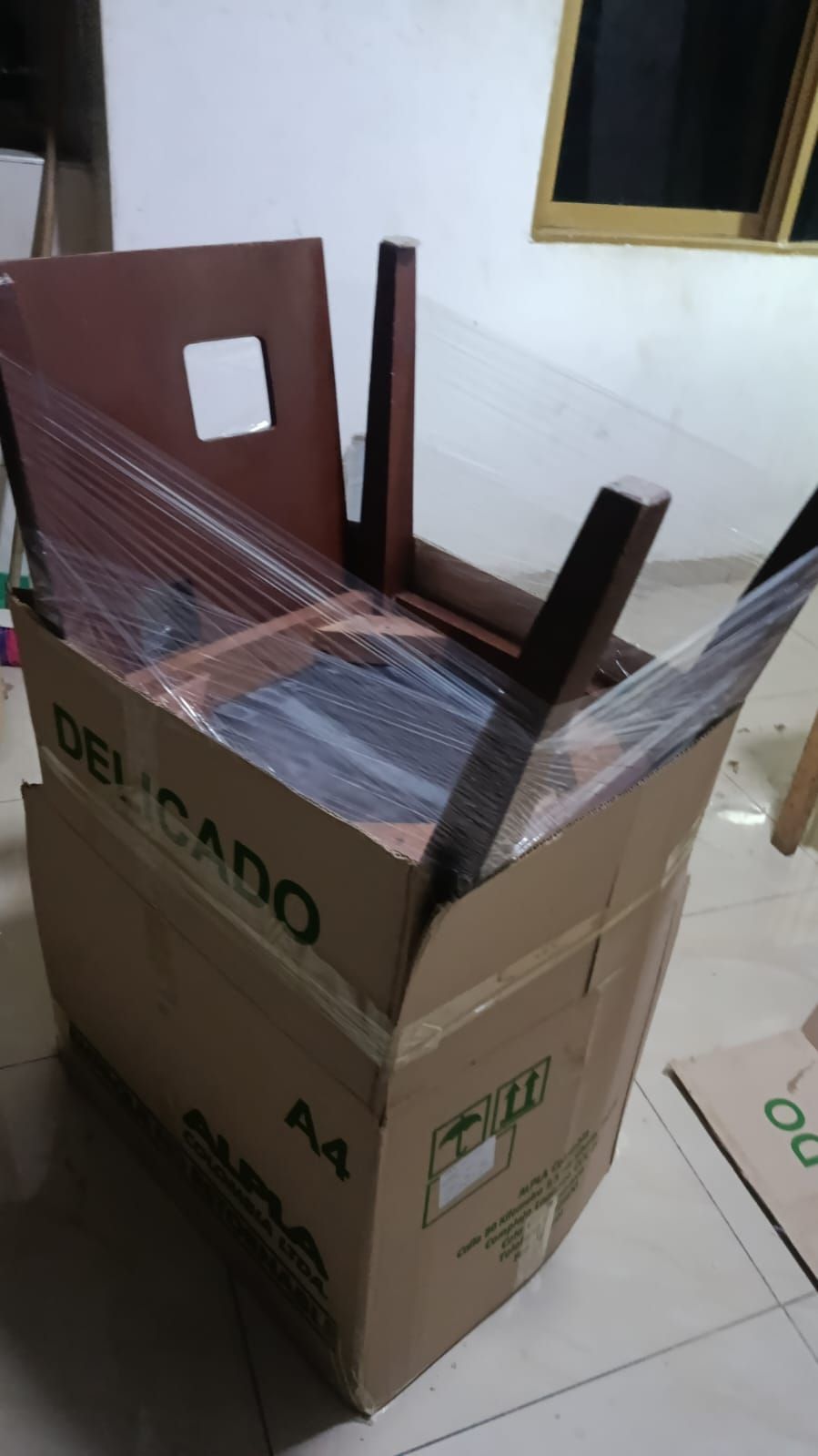 Caja de cartón embalada con una silla de madera marrón envuelta en plástico dentro de una habitación con azulejos.