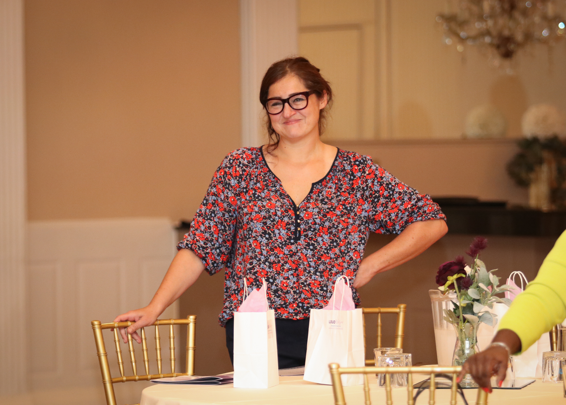 Woman with glasses, leaning on a gold chair at a table with gifts. She is wearing a floral top, in a well-lit room.
