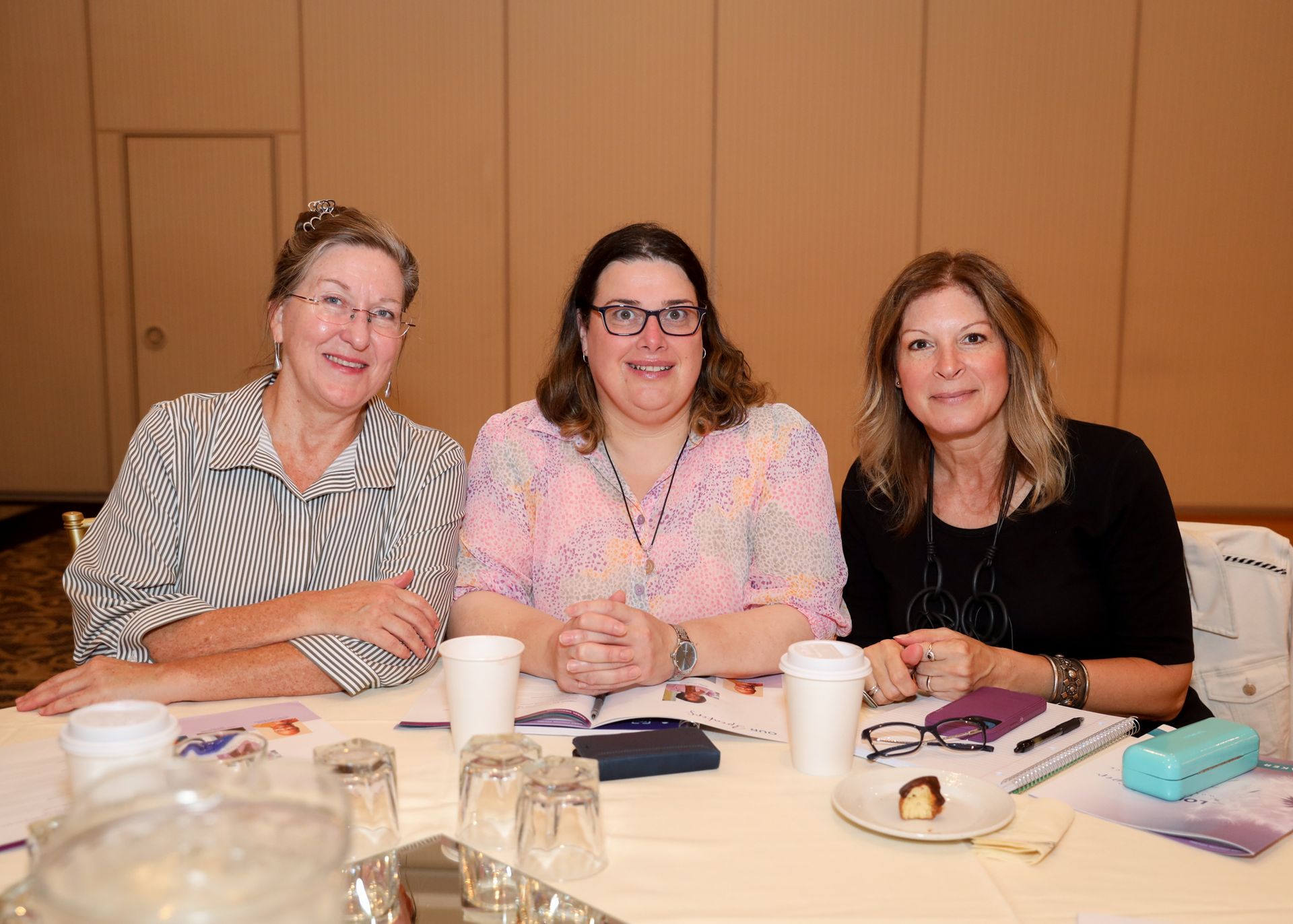 Three women sit at a table in a conference room, smiling. They have coffee cups, notebooks, and pens.