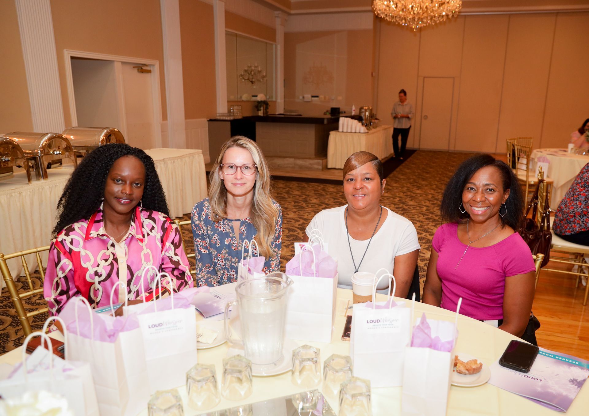 Four women sit around a table in a banquet hall, smiling. White gift bags and water glasses on the table.