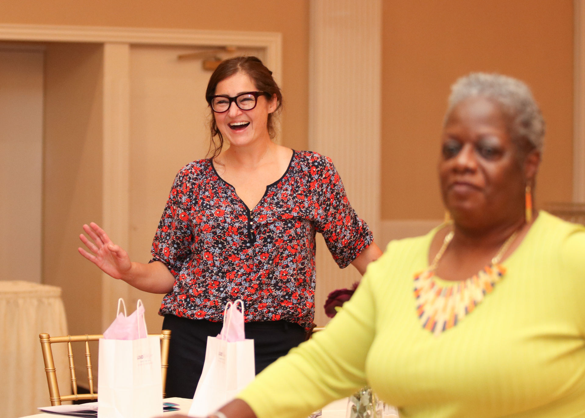 Woman with glasses laughs, arms raised, near a woman in yellow top at an event, near gift bags.