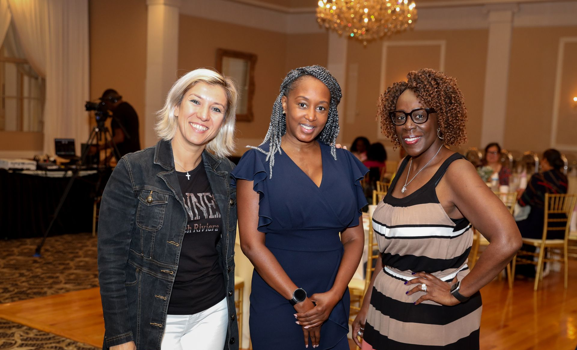 Three women pose smiling at a formal indoor event with chandeliers and round tables.