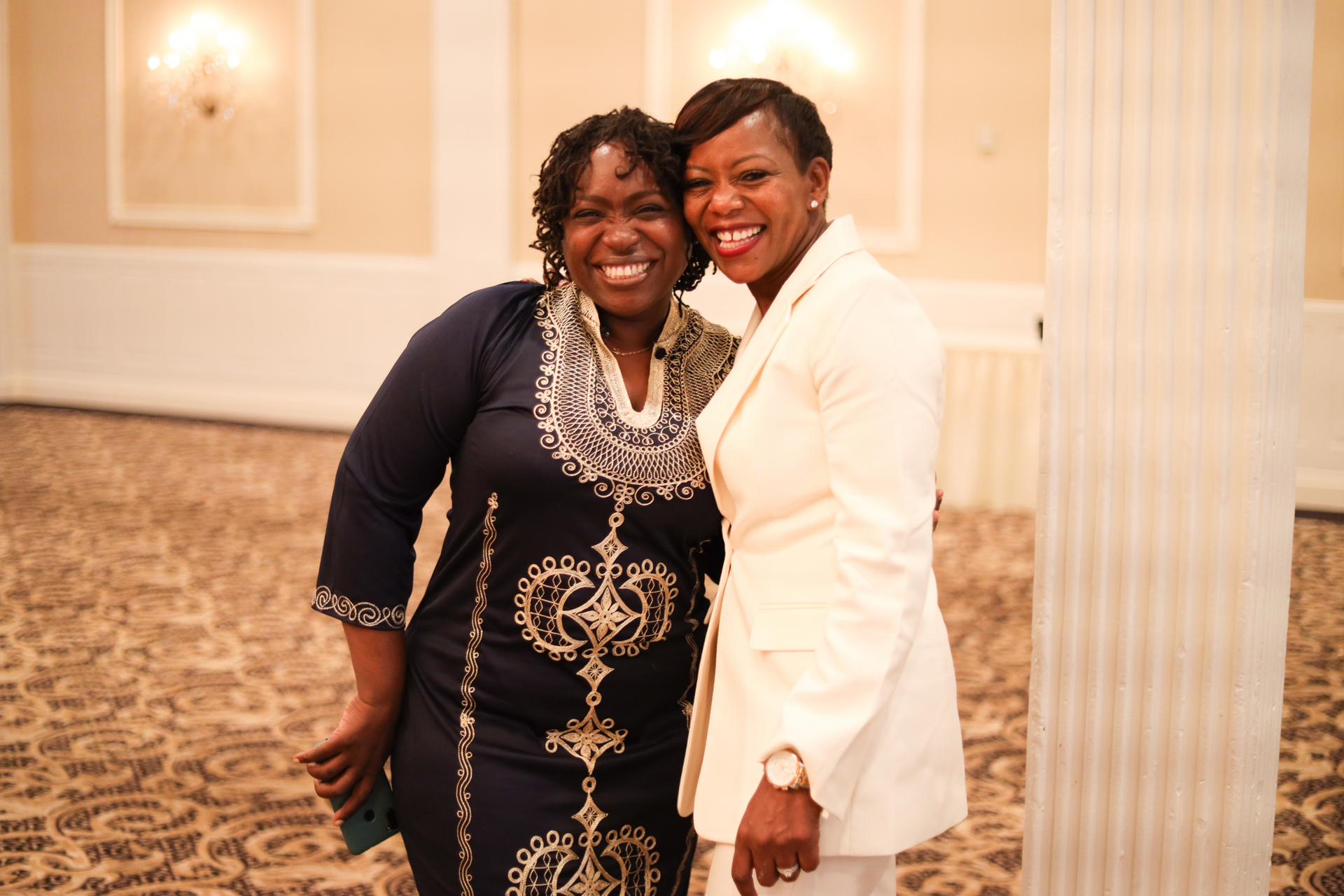 Two women smiling and posing for a photo in a well-lit room. One wears a blue patterned dress; the other a cream suit.