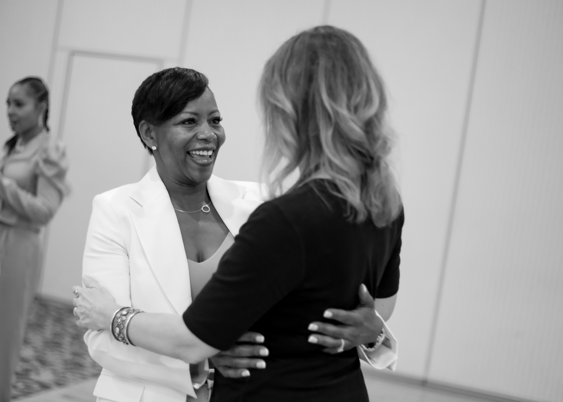 Two women embrace, smiling. One in white blazer, the other in black top. Woman in background watches.